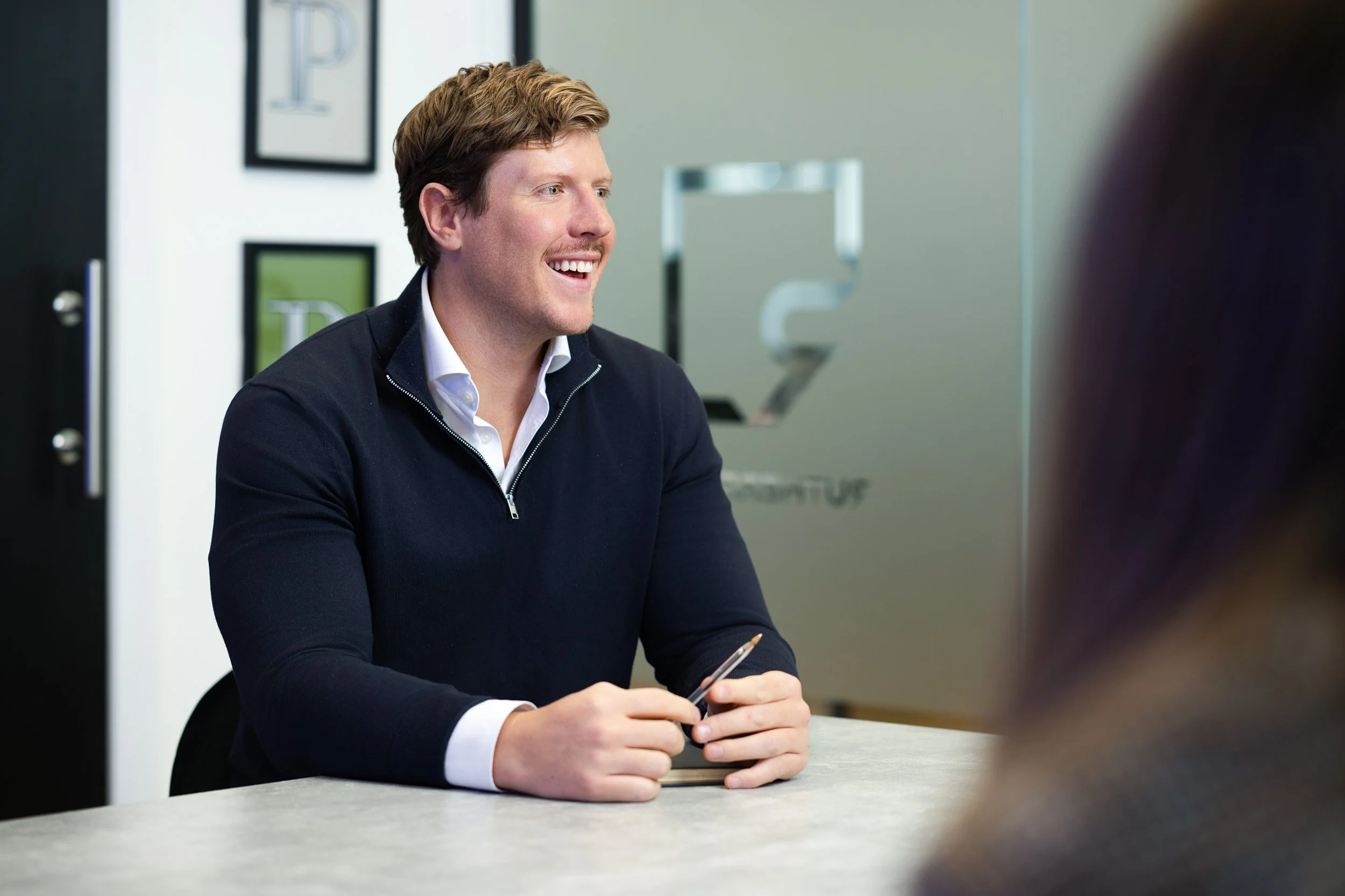 A man with light brown hair, wearing a white shirt and navy blue sweater, sitting at a desk in an office, smiling and holding a pen, engaged in conversation with someone off-camera.