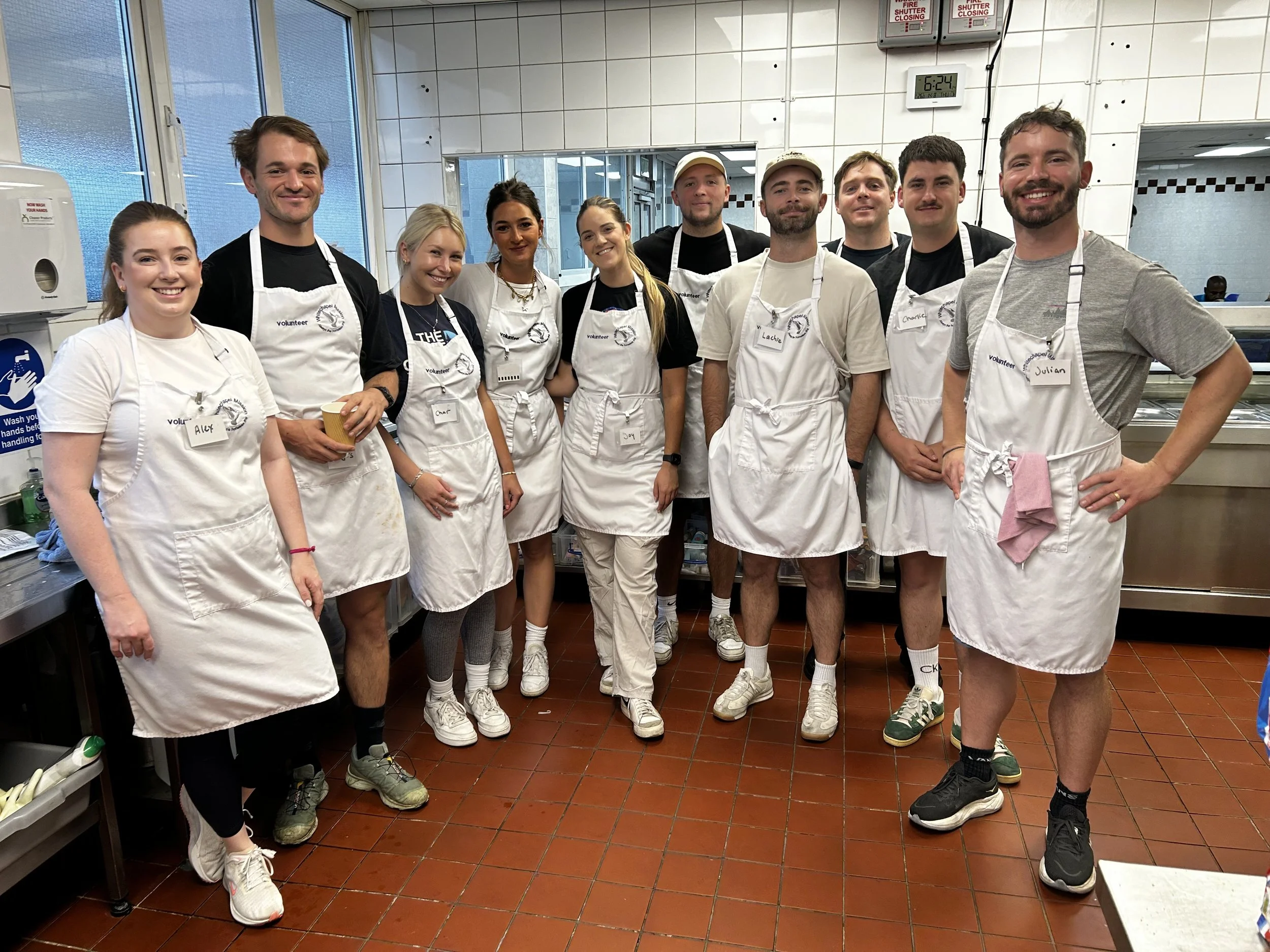 Group of people in a kitchen, wearing white aprons with name tags, some smiling and standing together.
