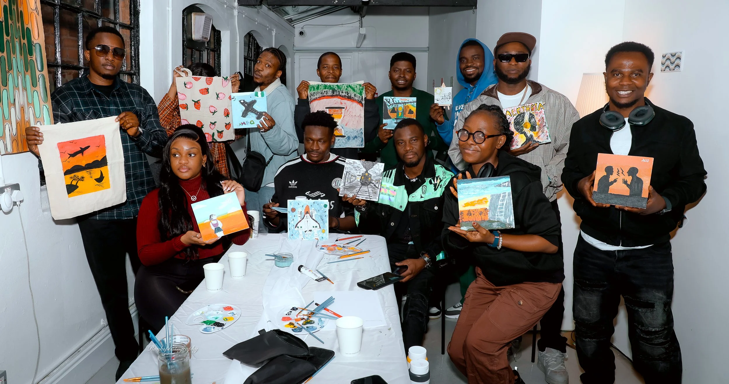 Group of people holding up their painted artworks in an indoor art workshop or gallery, with painting supplies on a table in front of them.