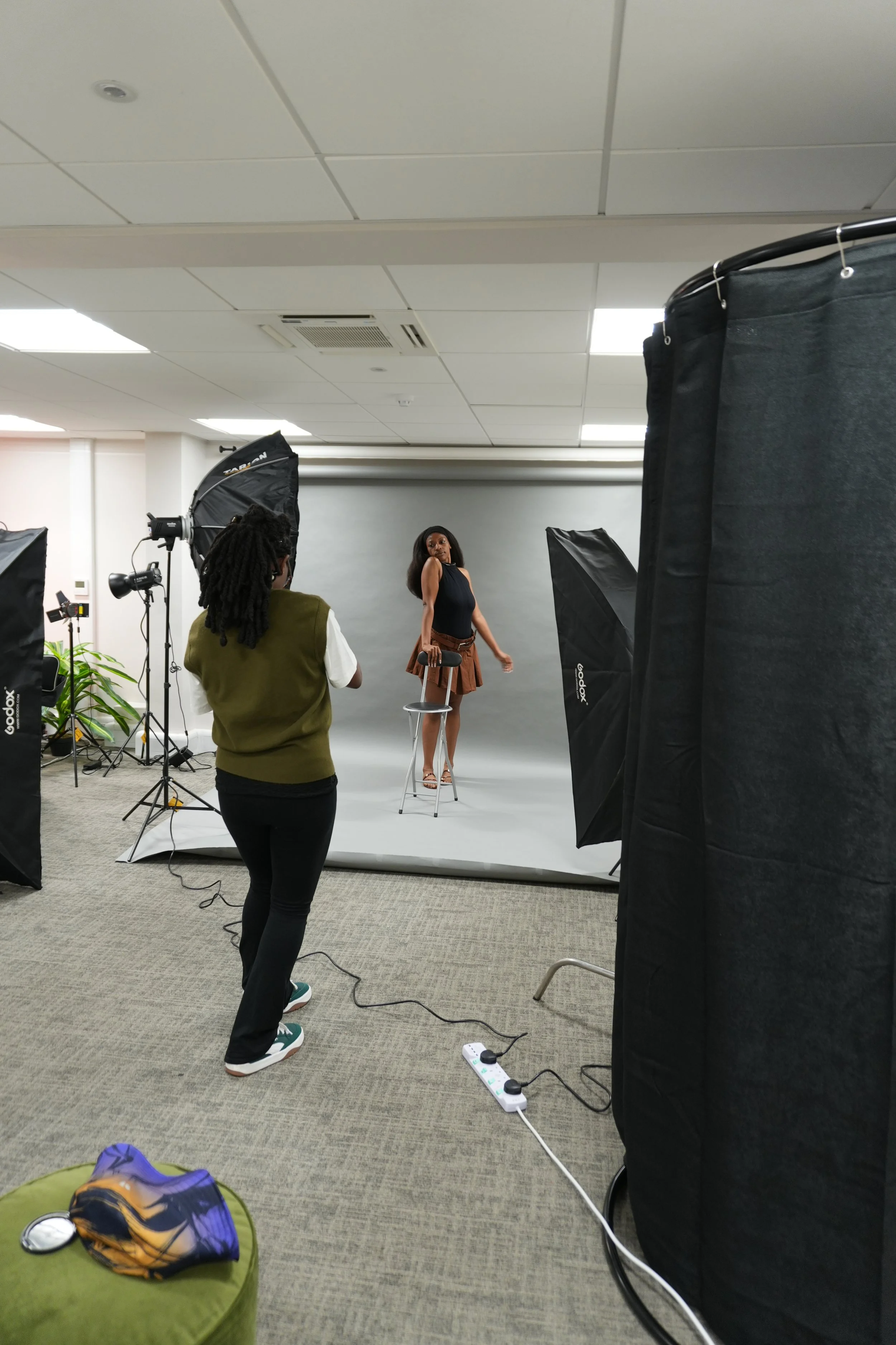 Photo of a woman standing in front of a gray backdrop while someone else takes her picture in a photography studio. The studio has softbox lights and various equipment, with a power strip on the carpeted floor.