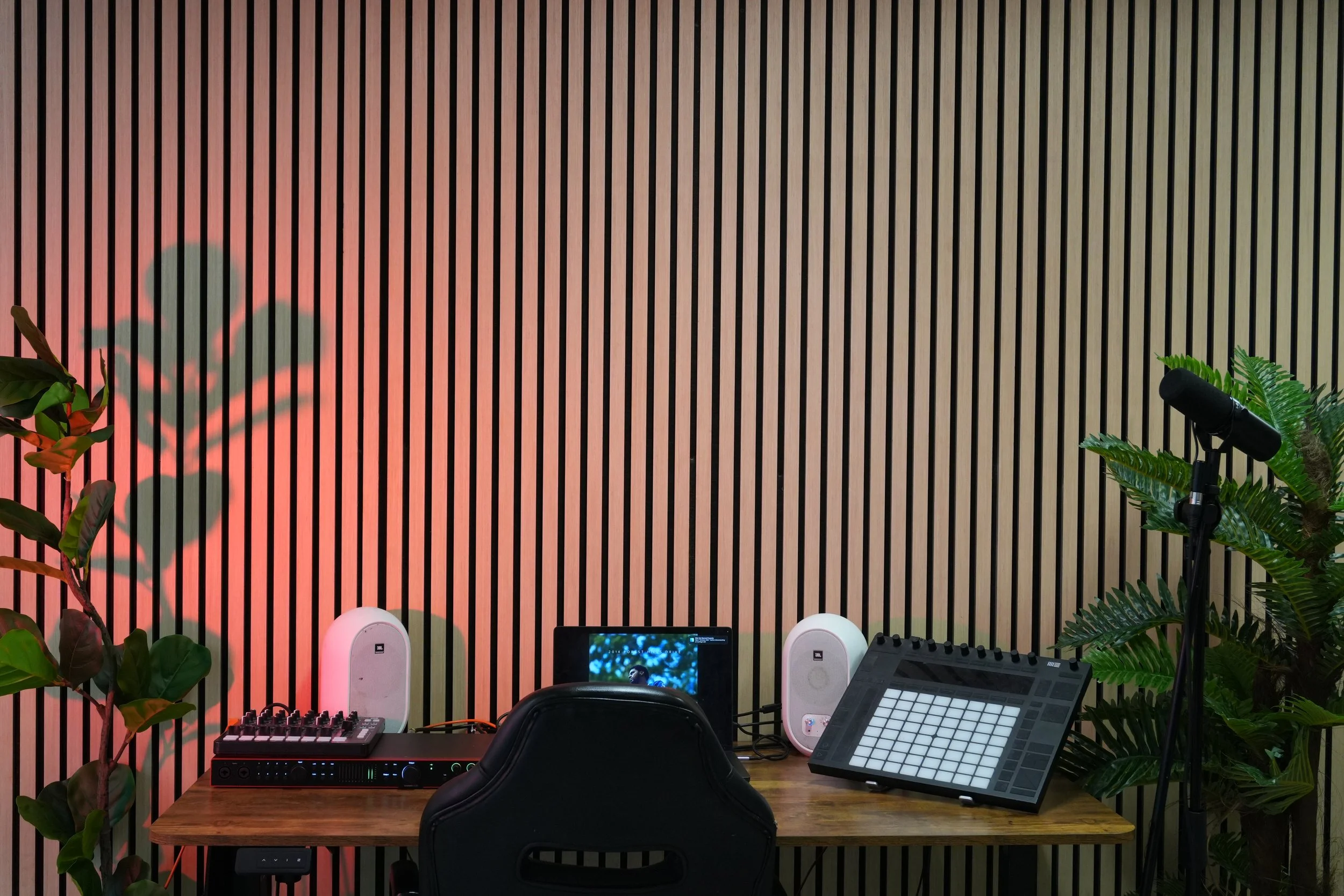 A music production workspace with a wooden table, two white speakers, a MIDI controller, a laptop, and a digital audio workstation on the screen, set against a vertically striped wooden wall with plants on both sides.