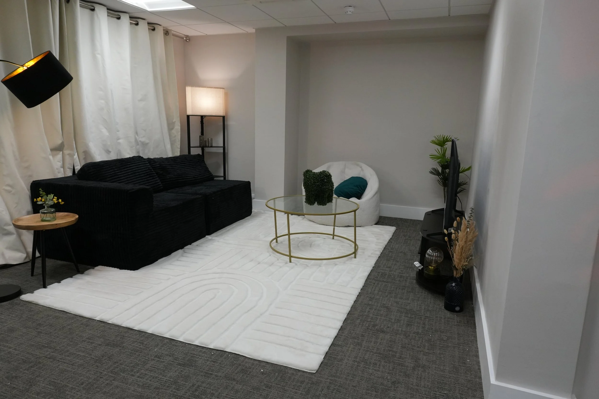 A modern living room with a black sofa, white curtains, a white rug, and a small coffee table with a green plant. There is a tall floor lamp and a bookshelf with candles. A white chair with a teal pillow is near a TV. Various decorative items and pla