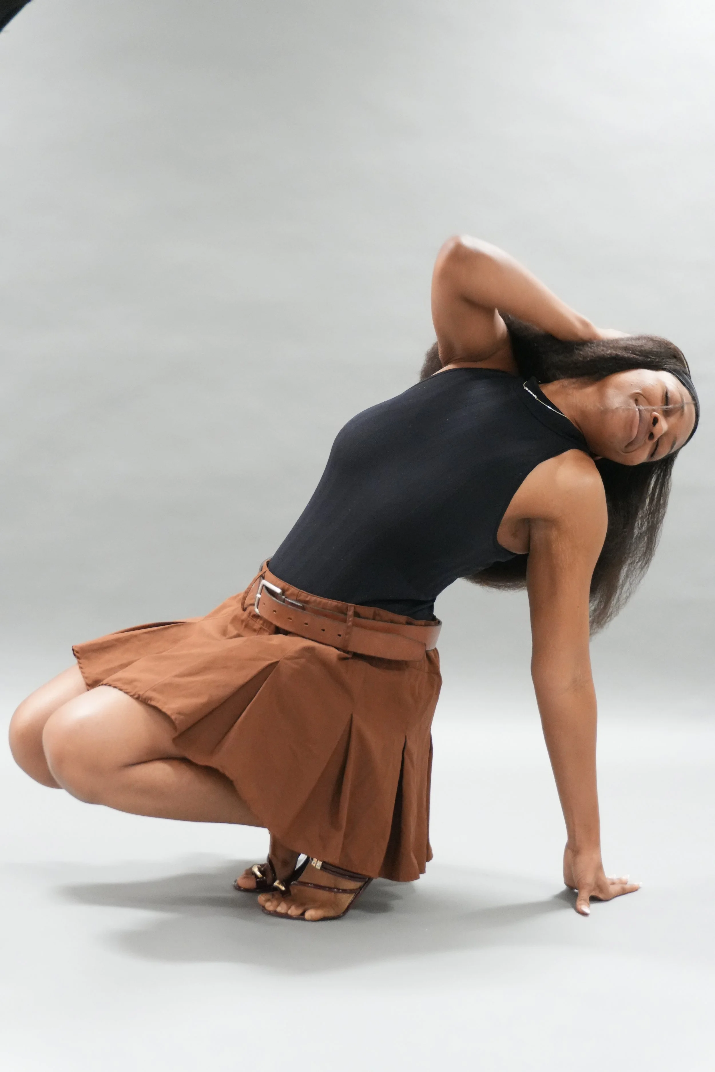 A woman in a black tank top and brown pleated skirt posing on the floor with one hand and knee on the ground, her other hand behind her head, and her long dark hair hanging down, against a plain light gray background.