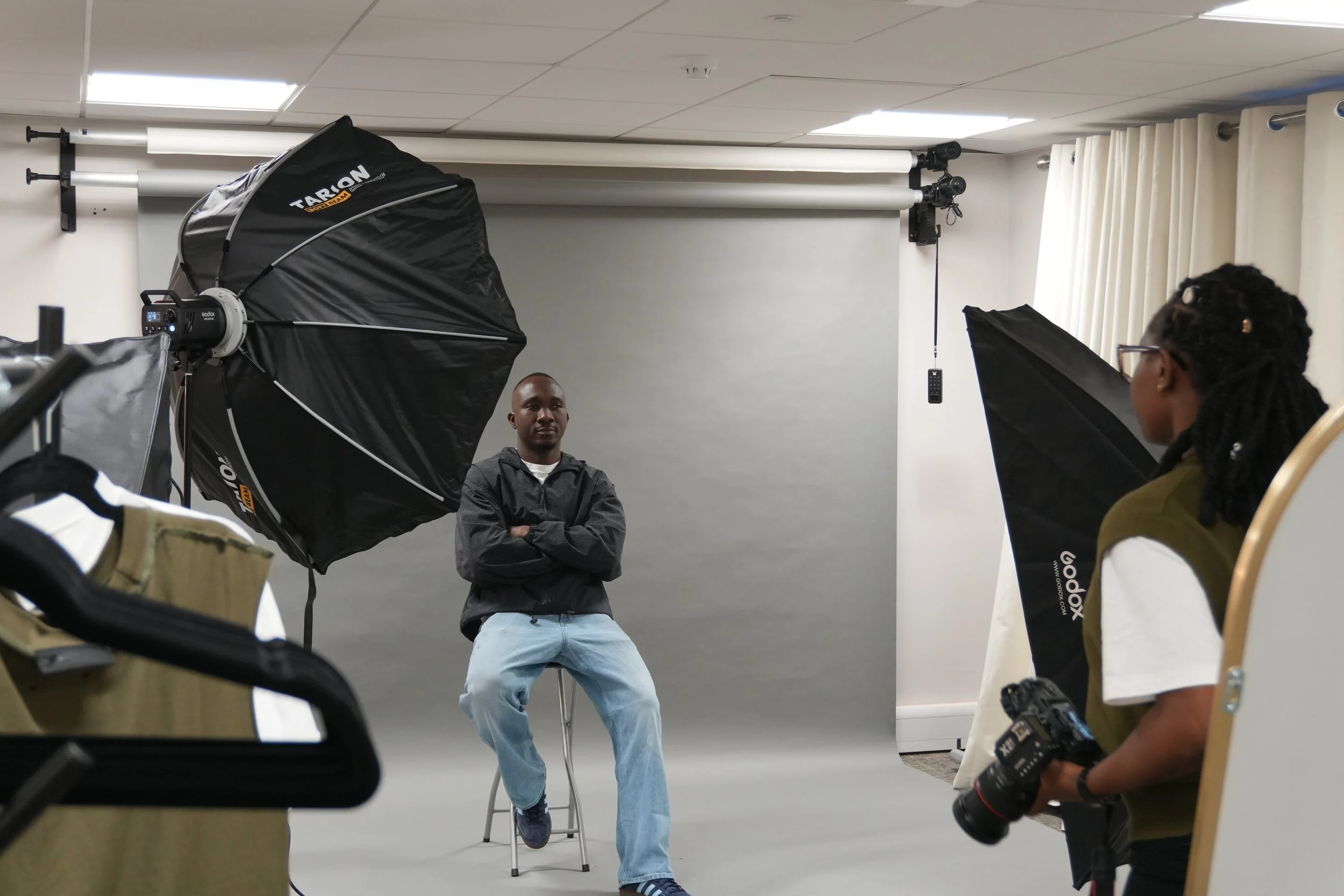 A photo studio with a man sitting on a stool, arms crossed, in front of a white backdrop, surrounded by professional lighting and equipment, with a woman taking photos.