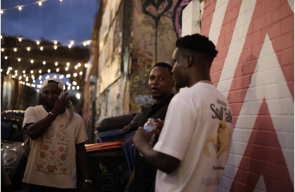Three young men having a conversation outdoors at night, with string lights overhead and colorful graffiti on the wall behind them.