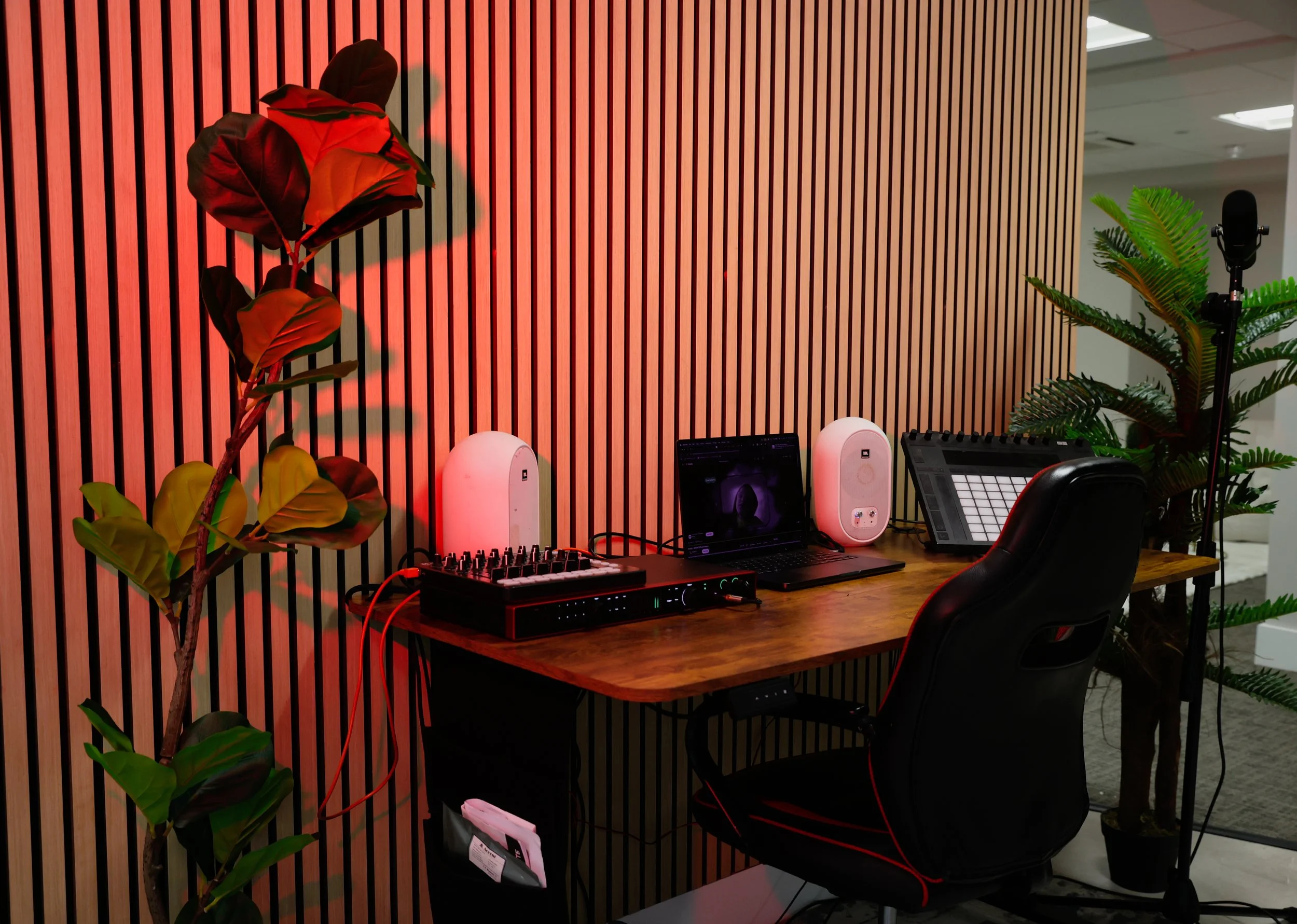 Office space with a wooden desk, two white speakers, a laptop, a music controller, headphones, and several green plants in the background and foreground.