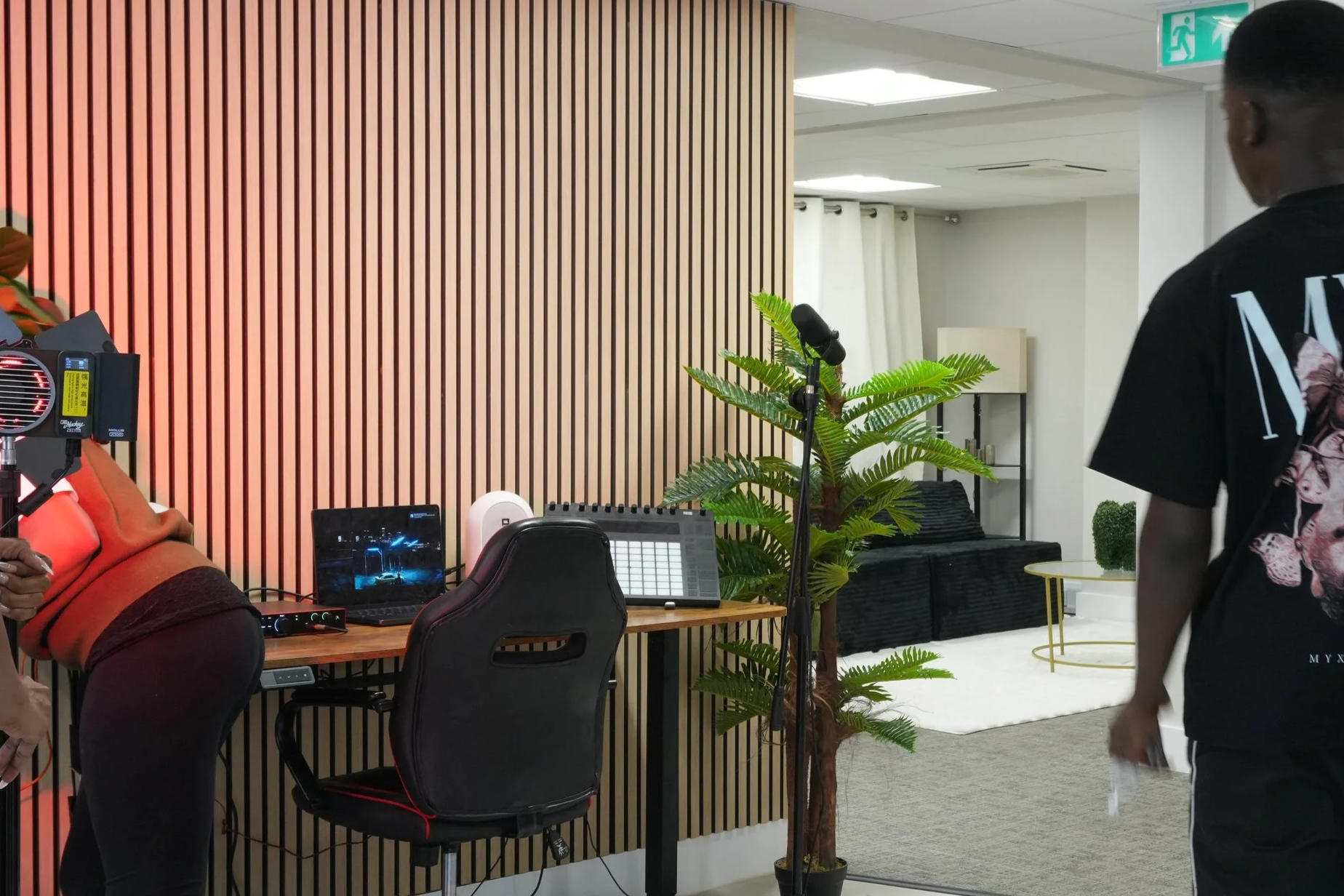 Interior of a modern office or studio with musical equipment, including a microphone, laptop, and audio interface, alongside a wooden desk and black chair, and a large green plant in front of a wooden slat wall.