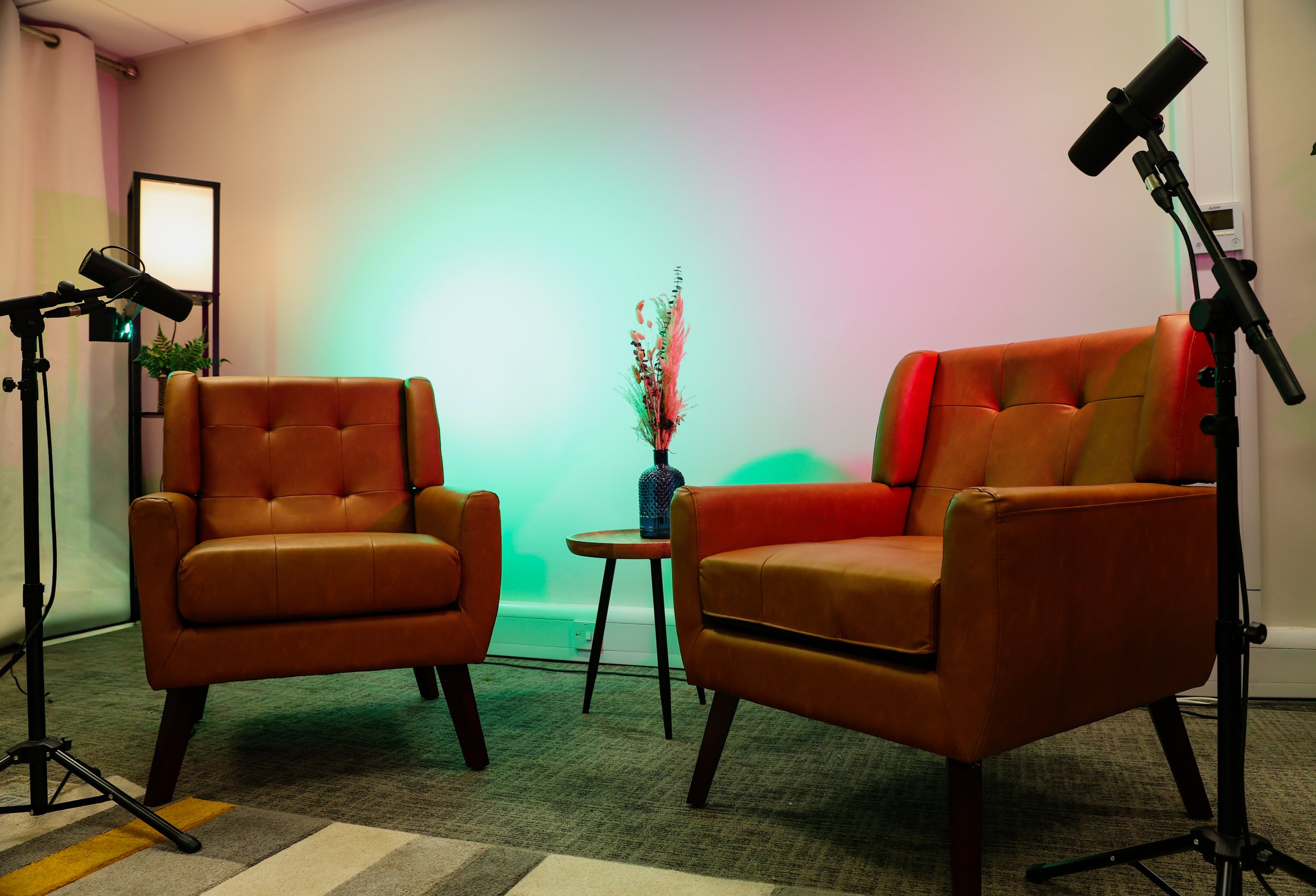 A cozy backstage area with two vintage brown leather armchairs, a small round wooden table with a decorative vase of pink and beige flowers, a potted fern, surrounded by photography lighting equipment, and a pastel-colored wall in the background.