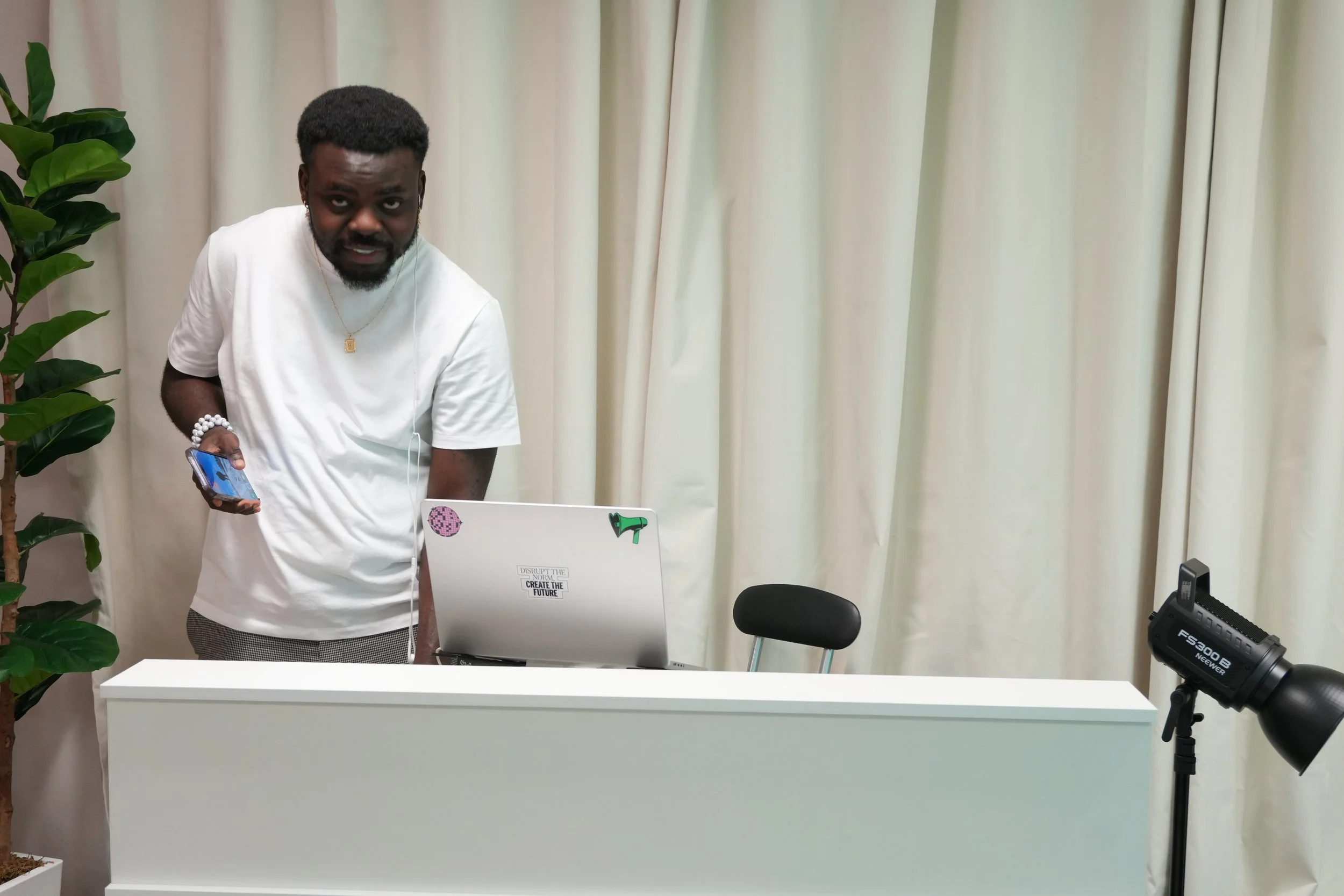 A man with dark hair and a beard wearing a white t-shirt, holding a smartphone in his right hand, standing behind a white desk with a laptop and a sticker that says "Disrupt the Norm, Create the Future," in a room with beige curtains, a green plant, 