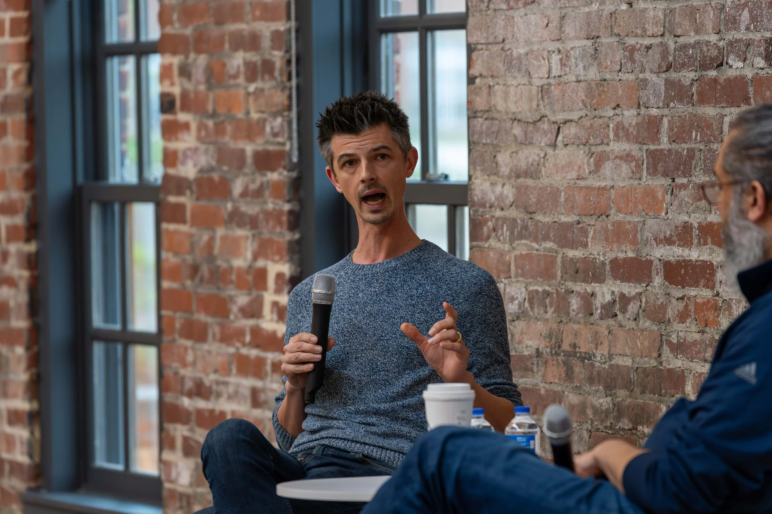 Man speaking into a microphone during a discussion, sitting next to a person with a beard, glasses, and gray hair, in a room with brick walls and large windows.