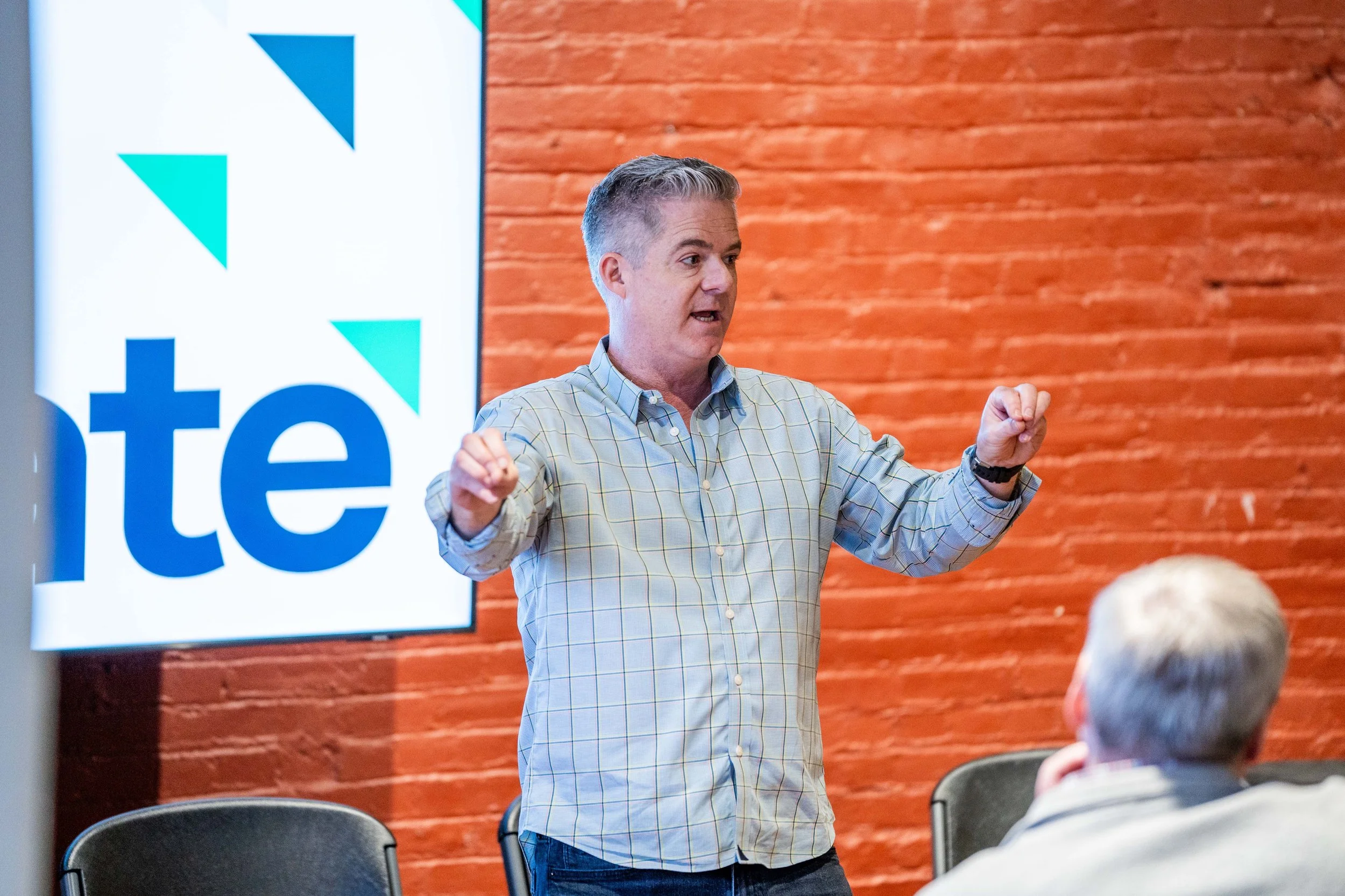 A man giving a presentation or speech in front of a screen with a partial logo on a brick wall background, with a seated audience member in the foreground.