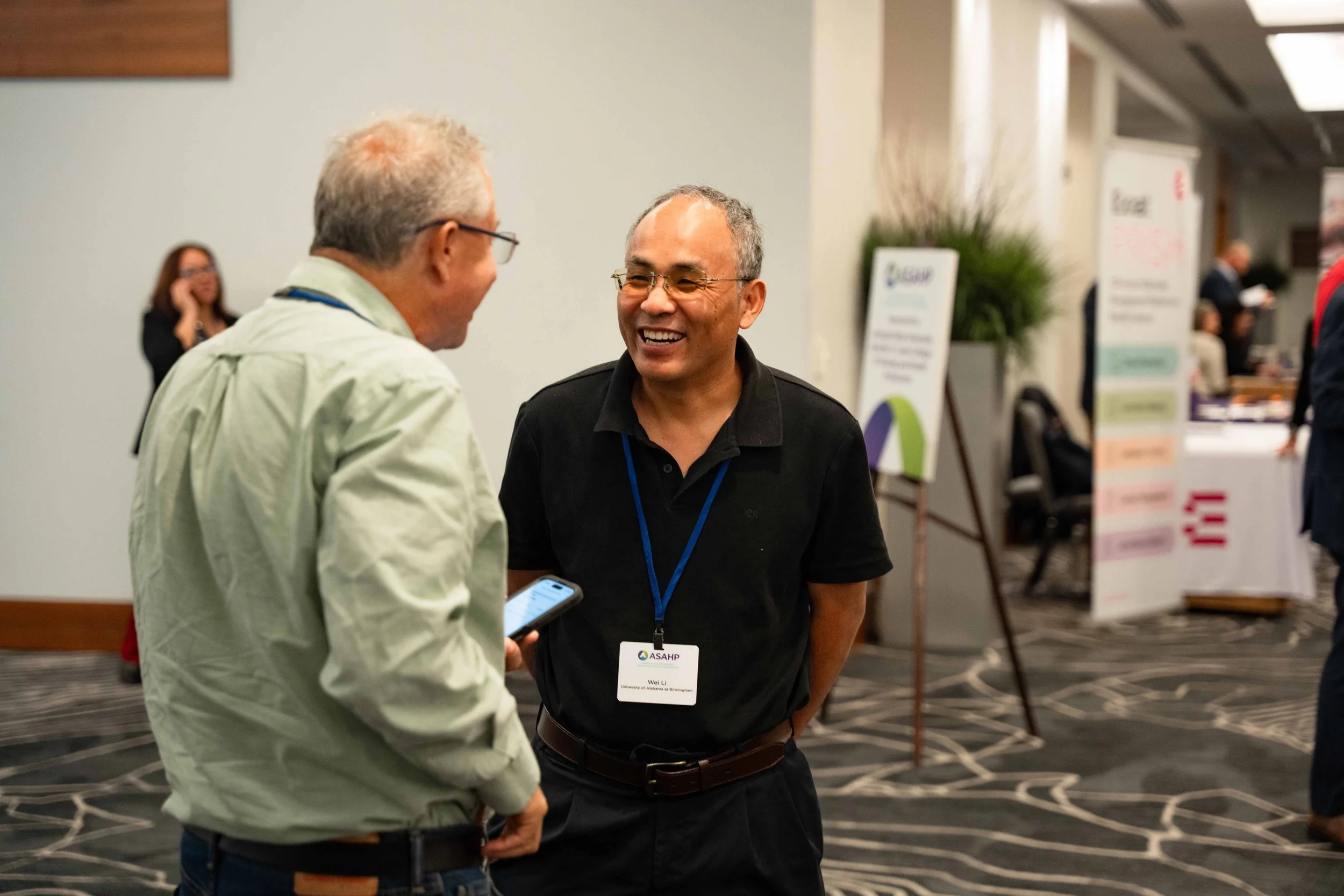 Two men are talking and smiling at a conference. One has gray hair and glasses, holding a phone, and the other is wearing a black polo shirt with a conference badge. In the background, there are banners, other people, and conference booths.