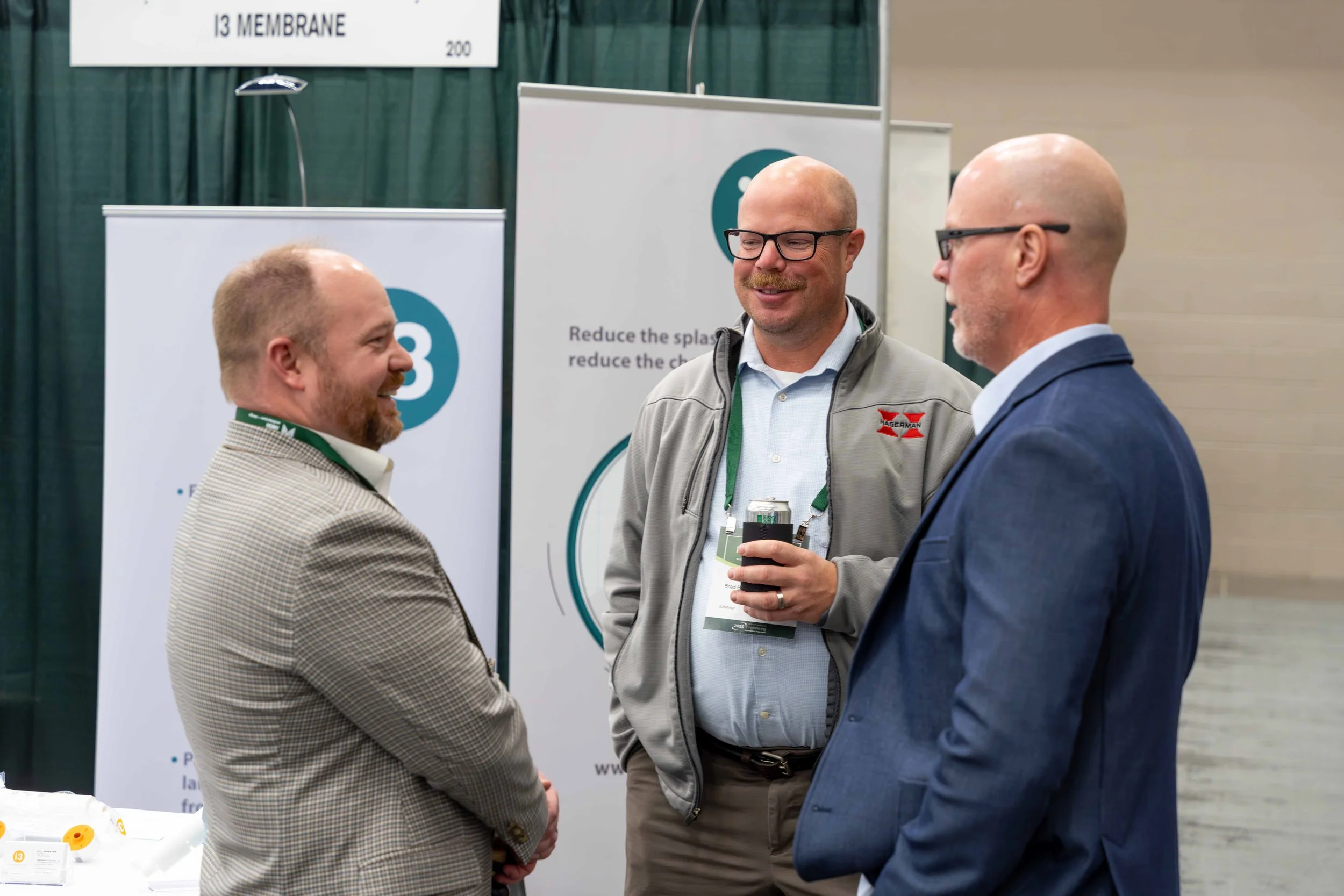 Three men in business attire and casual clothing are engaged in conversation at a conference or trade show. They are smiling and holding drinks, with a booth in the background featuring banners and a green curtain.