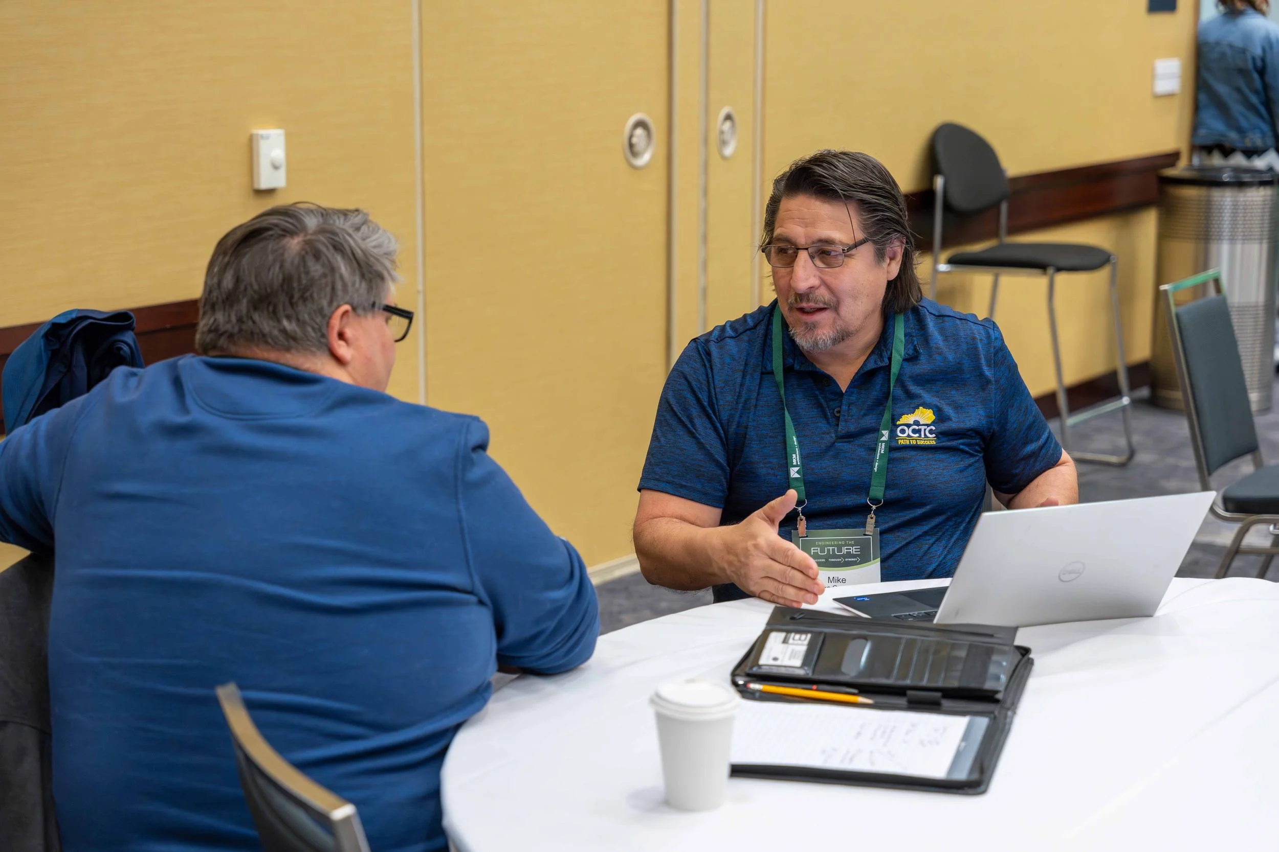 Two men sitting at a round table having a discussion indoors. One man is facing away from the camera, wearing glasses and a blue jacket. The other man, with longer hair and a beard, is facing the camera, wearing a blue shirt with a logo and an ID bad