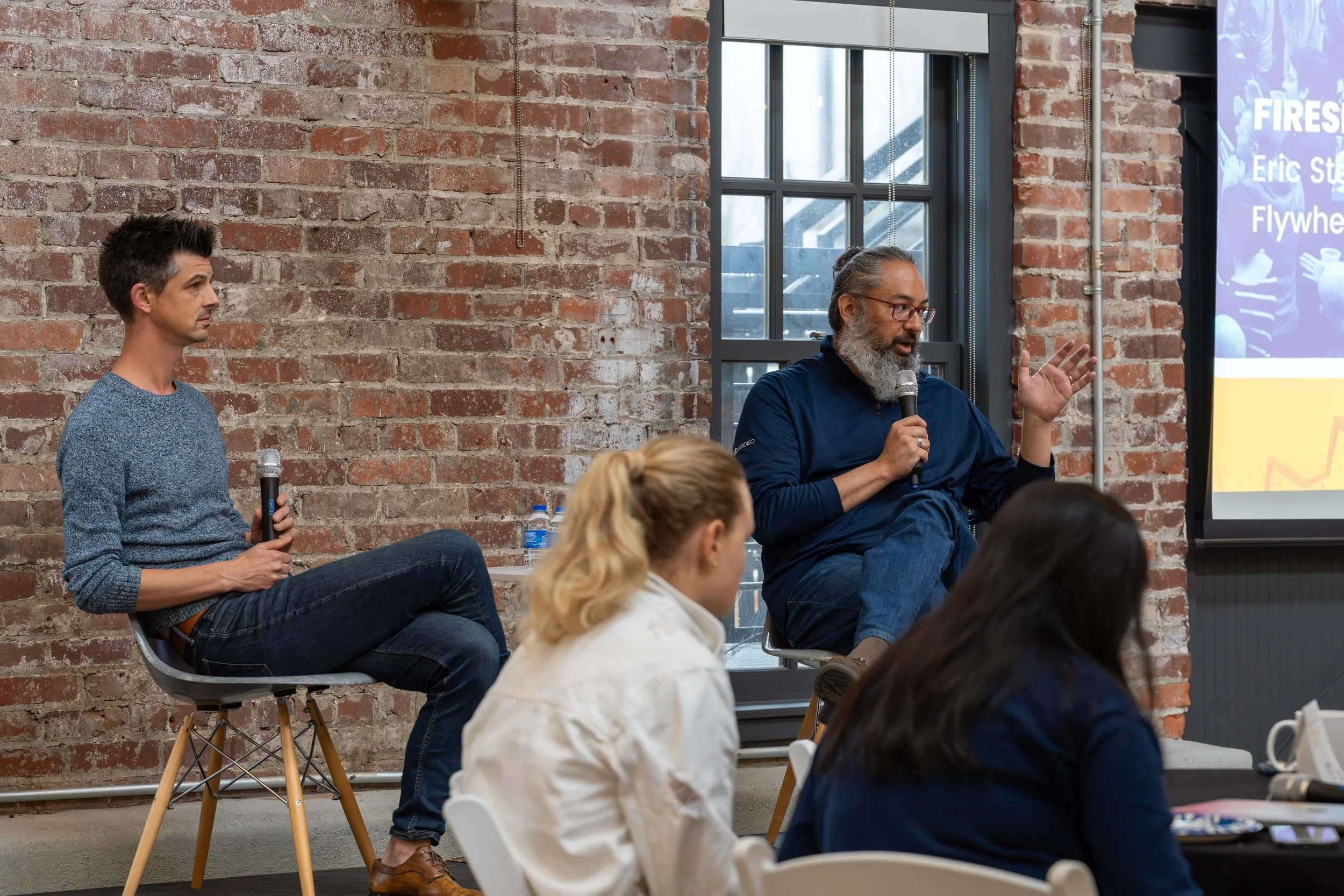 A man with gray hair and beard wearing glasses and a navy jacket, holding a microphone, speaking during a panel discussion in a brick-walled room with a large screen displaying a presentation slide.