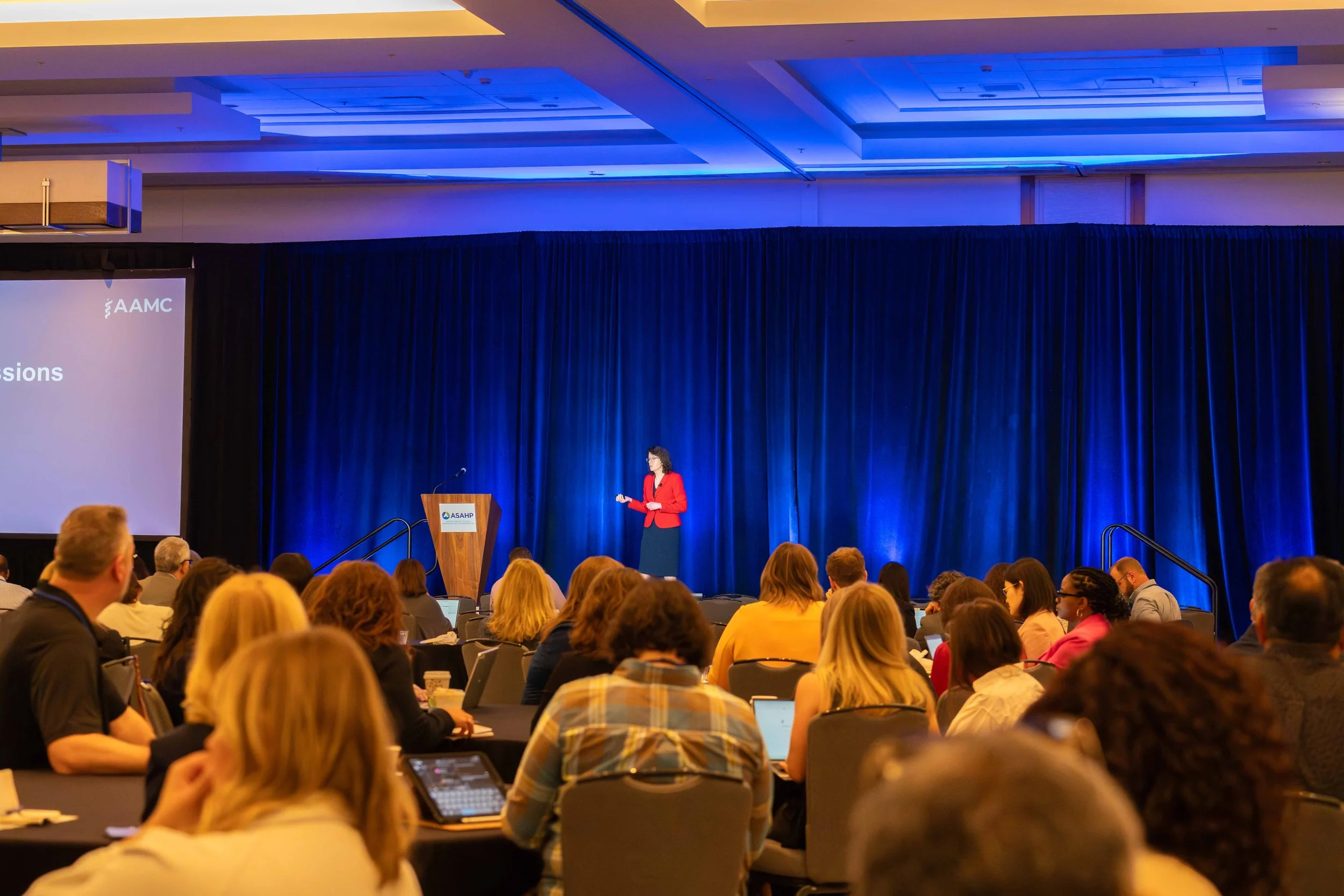 A woman in a red blazer giving a presentation on stage at a conference, with an audience seated at tables watching.
