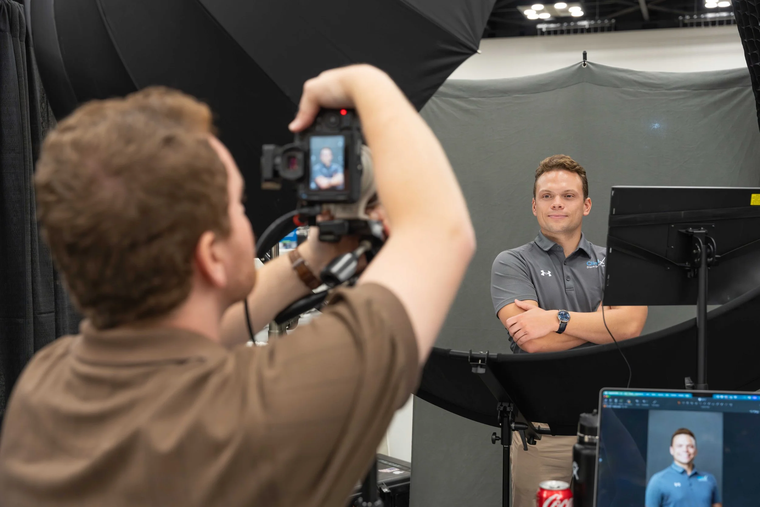 A man in a gray polo shirt with a logo on it is posing with arms crossed in front of a camera, being photographed or filmed by another man with a camera at a professional photo shoot. There is a computer monitor displaying an image of the man in the background.