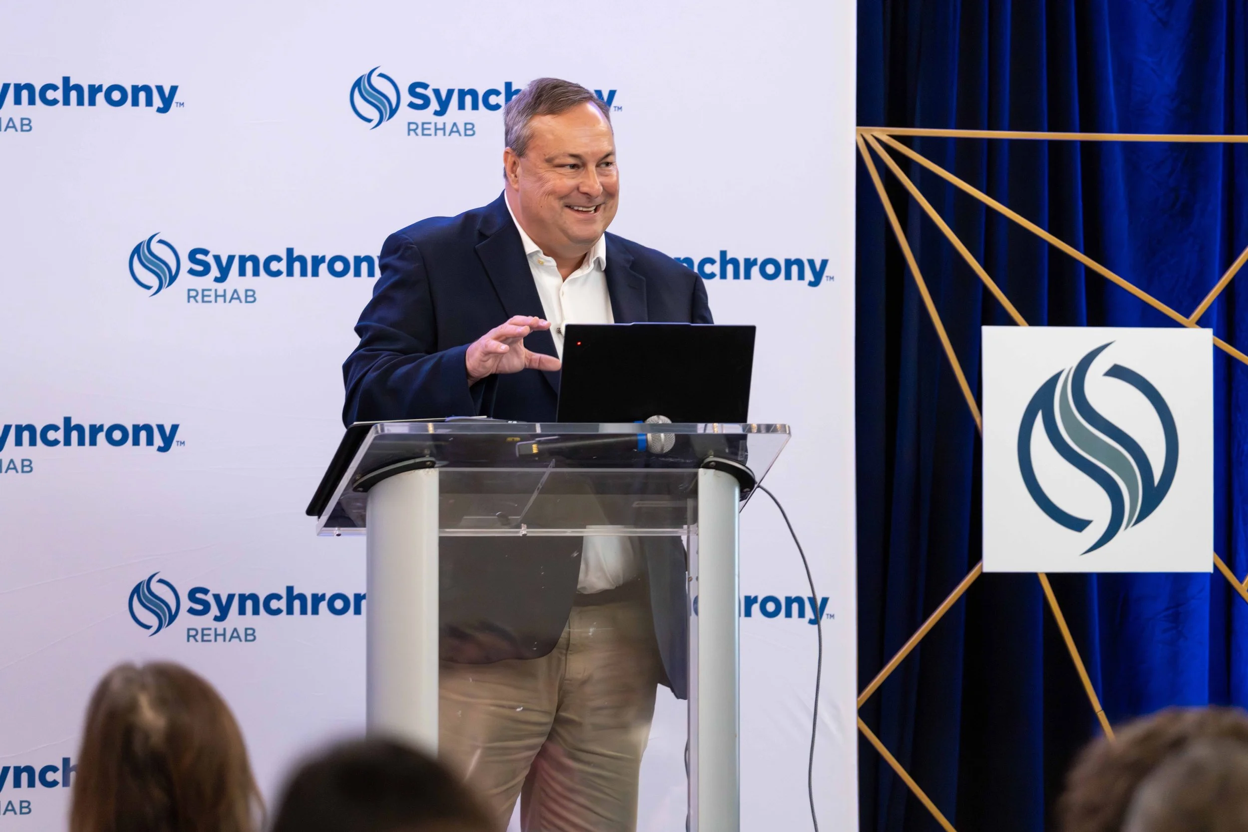 A man in a dark blazer and white shirt gesturing while standing at a clear podium with a laptop, in front of a backdrop with the Synchrony Rehab logo. The background includes a patterned curtain and the Synchrony logo on the side.