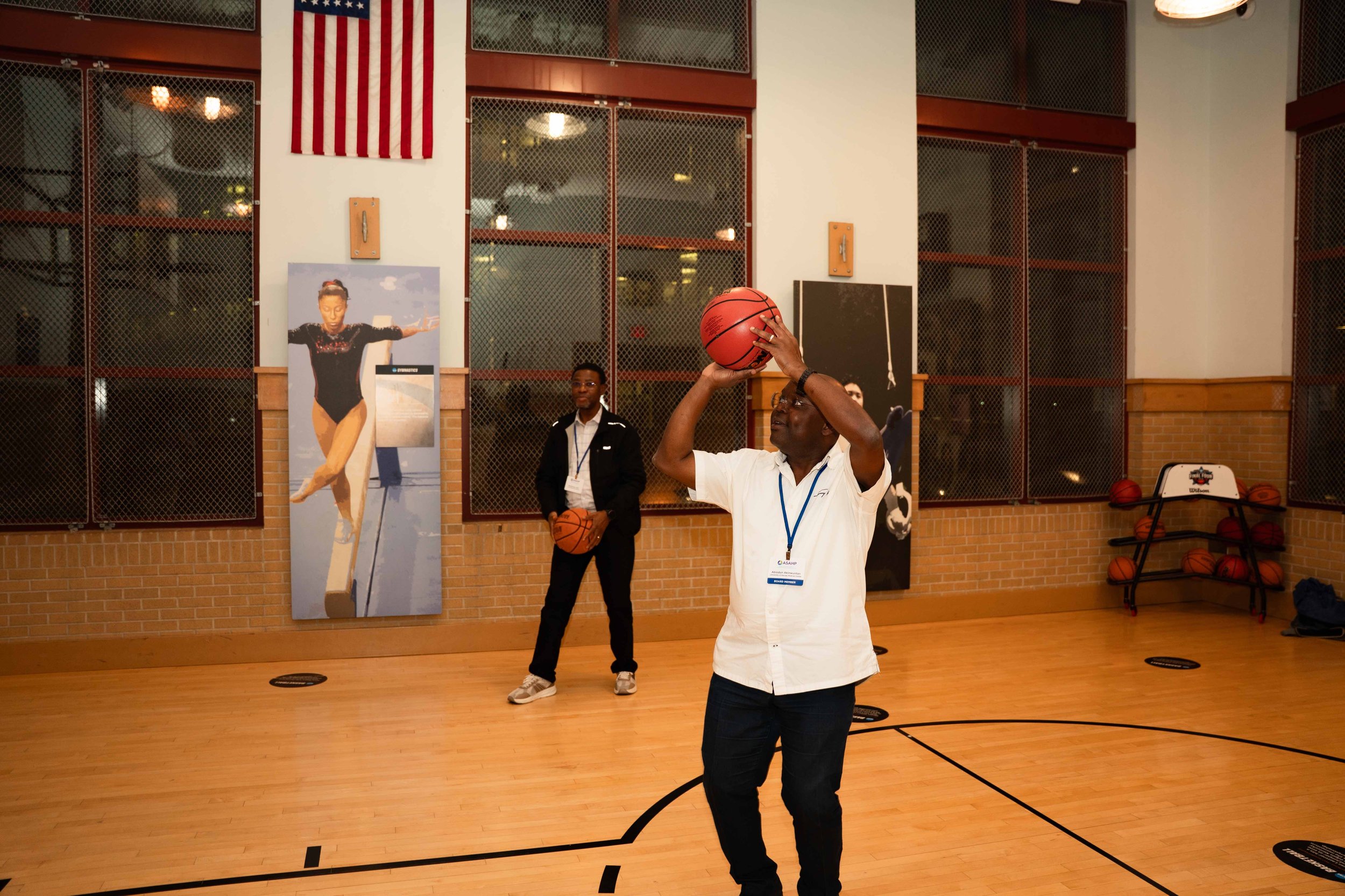 A man standing in a gymnasium preparing to shoot a basketball, with another person holding a basketball in the background, and a basketball rack with multiple balls. The gym has brick walls, large windows, and a mural of a gymnast on the wall.