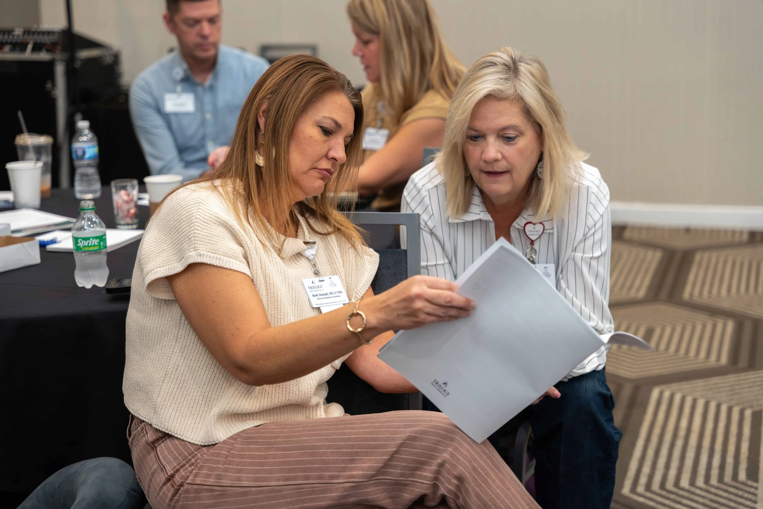 Two women engaged in a discussion over documents during a meeting, with other attendees and items like water bottles and coffee cups on the table in the background.