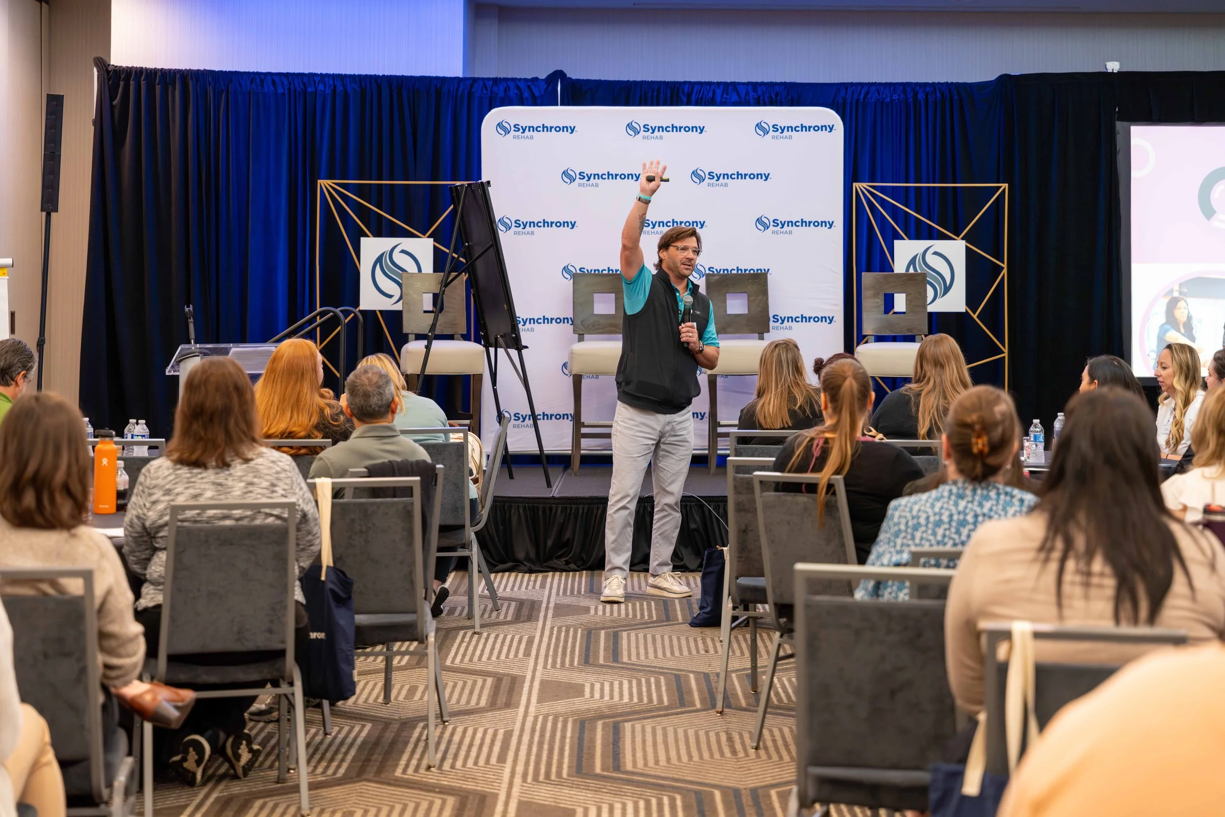 A man speaking on stage at a conference with a blue curtain backdrop and banners that read 'Synchrony Rehab,' audience seated in rows, some taking notes, with a large projection screen to the right.