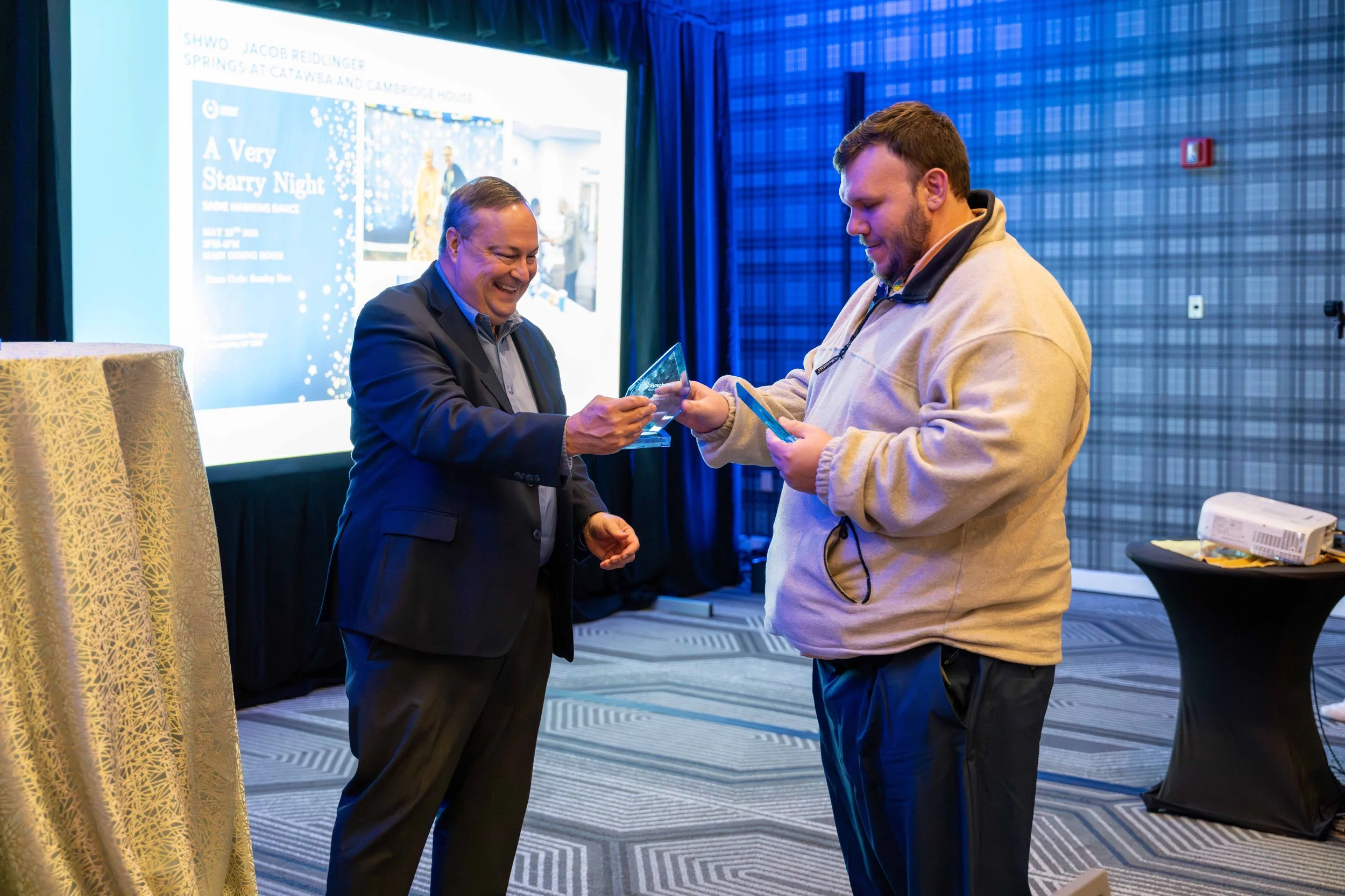 Two men exchanging awards at an indoor event, with a presentation slide about a spring event in the background.