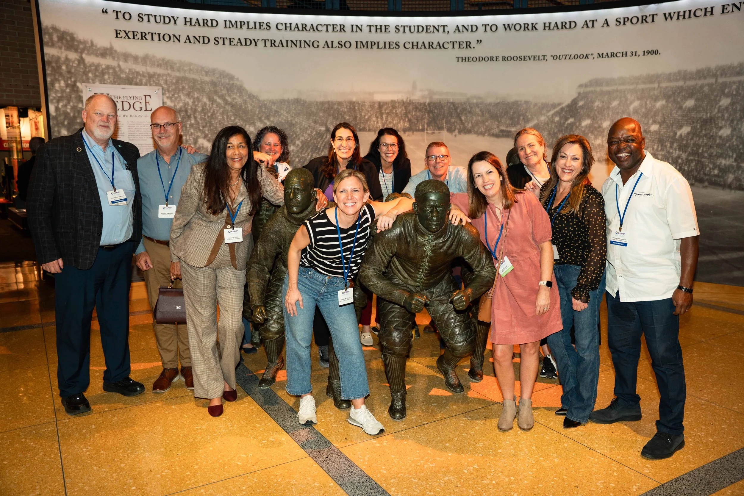 Group of diverse people smiling and posing behind bronze statues of sports players at an indoor venue. A quote about studying and character is visible on the wall behind them.