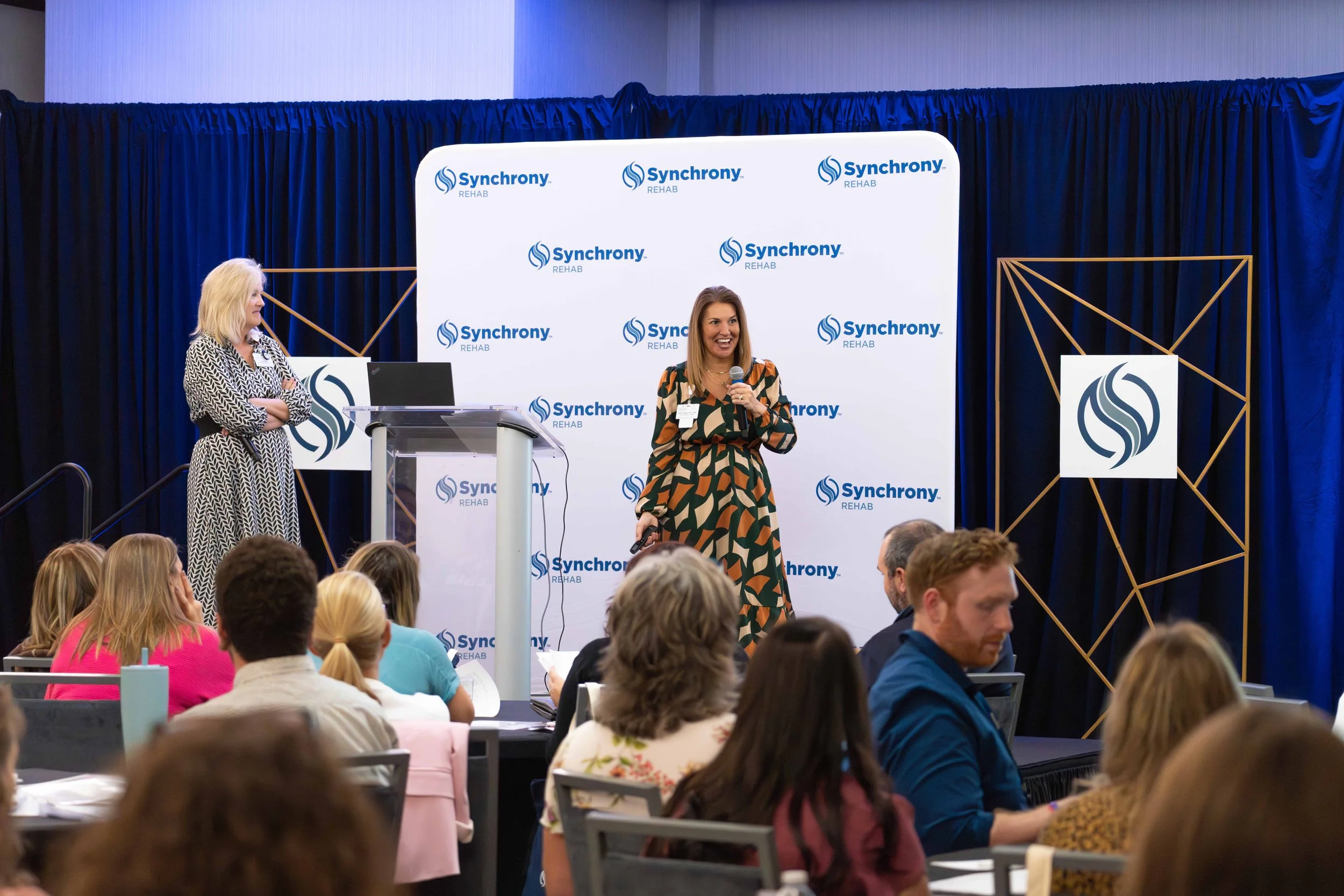 A woman in a colorful patterned dress speaking into a microphone on a stage at a Synchrony Rehab conference, with a large backdrop featuring the Synchrony logo behind her. Another woman stands to the side of the stage near a podium with a laptop, and