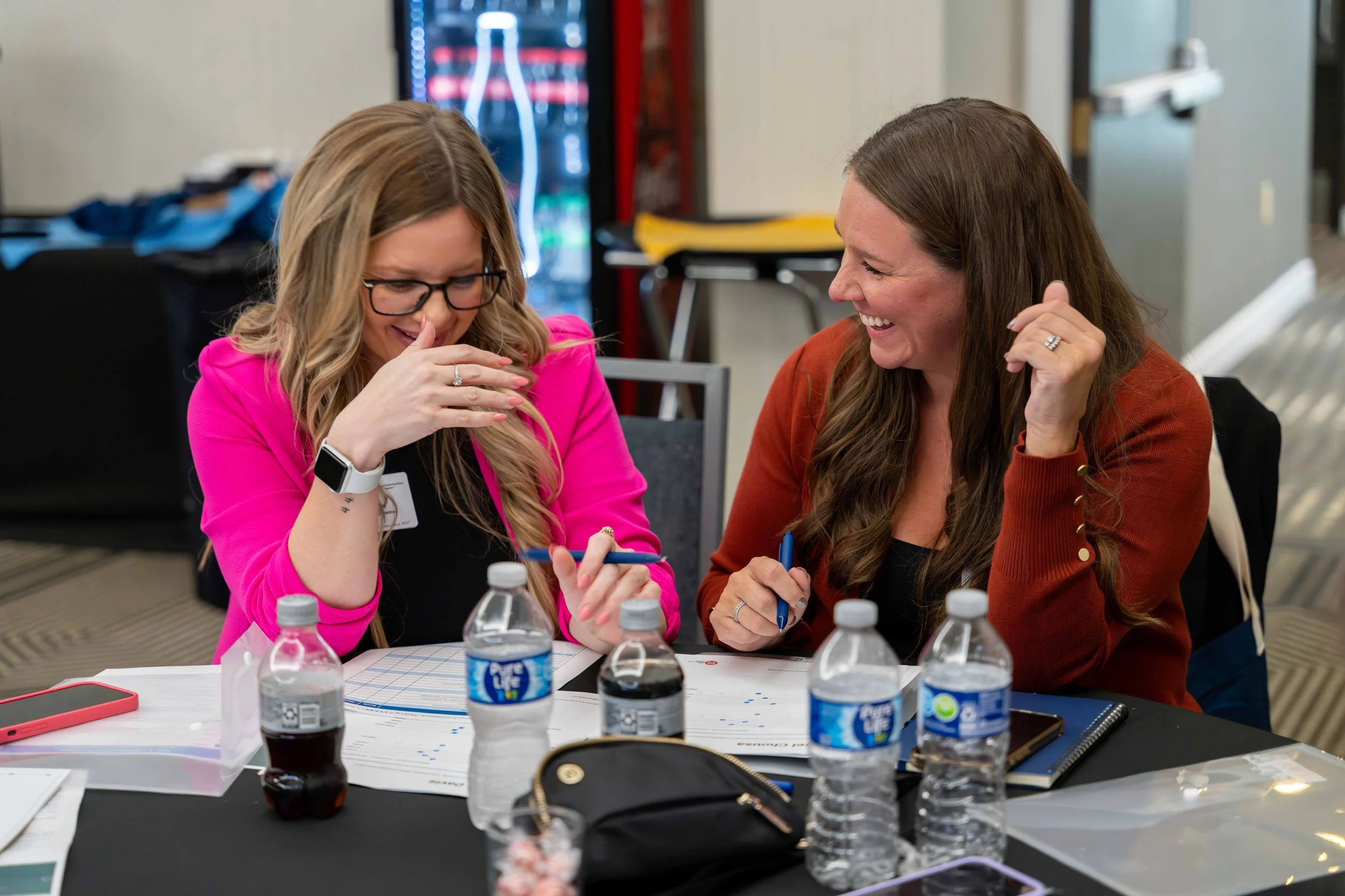 Two women sit at a table, laughing and smiling, surrounded by water bottles, papers, and personal items.