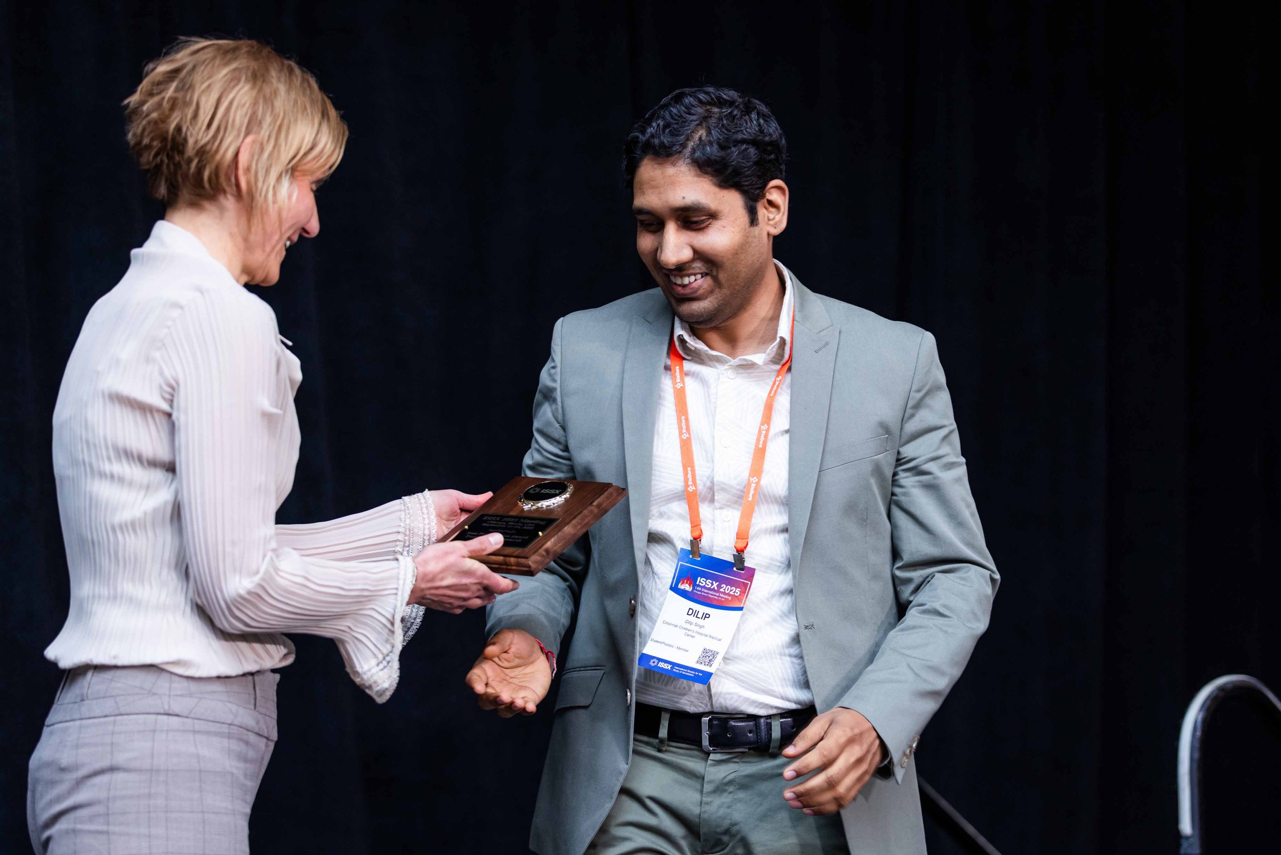 A woman presenting an award to a man at a conference or event. The woman is showing the award plaque to the man, who is smiling. Both are dressed formally, and the woman is wearing a light-colored striped shirt while the man is wearing a gray suit.