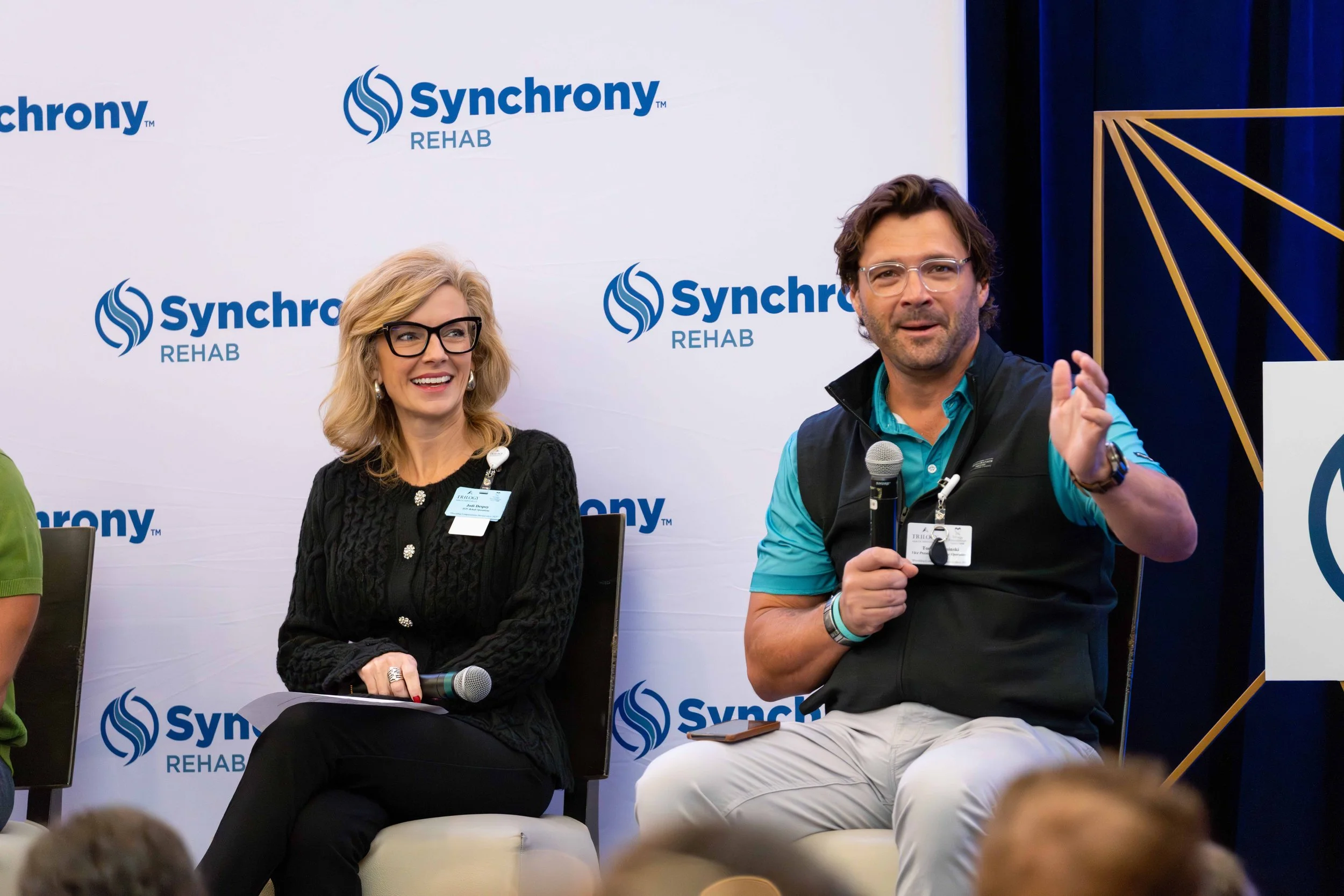 Two panelists, a woman with blonde hair and glasses, and a man with dark hair and glasses, are seated on stage at a conference with a backdrop displaying the logo of Synchrony Rehab. The man is holding a microphone and appears to be speaking, while t