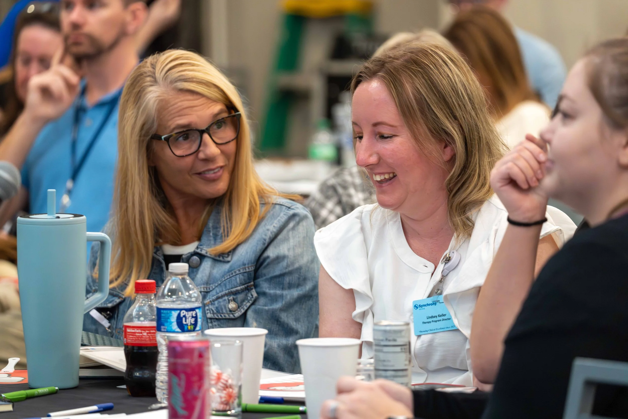 Three women at a table engaged in a lively conversation, with various drinks and objects on the table, in a busy indoor setting.