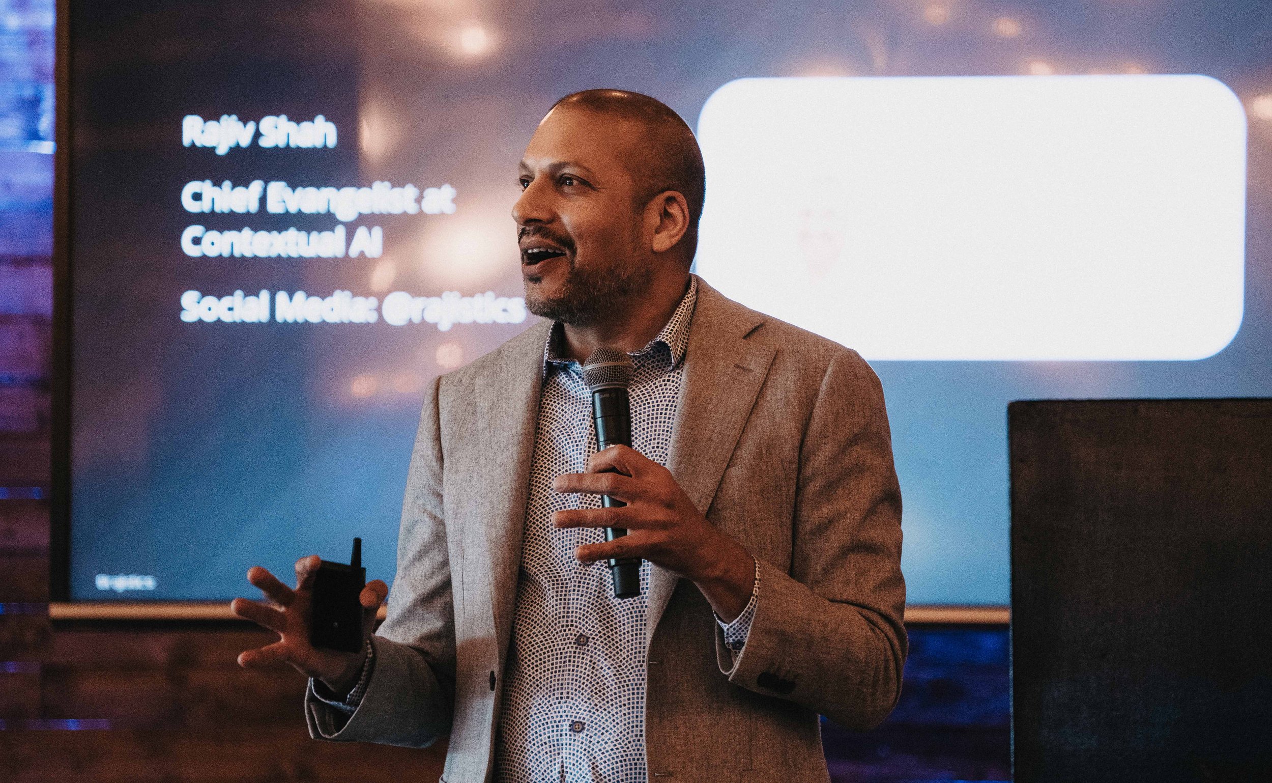 A man in a beige blazer and patterned shirt giving a presentation with a microphone in front of a screen that displays his name and title, "Rejv Shah, Chief Evangelist at Conversant AI, Social Media Oracles."