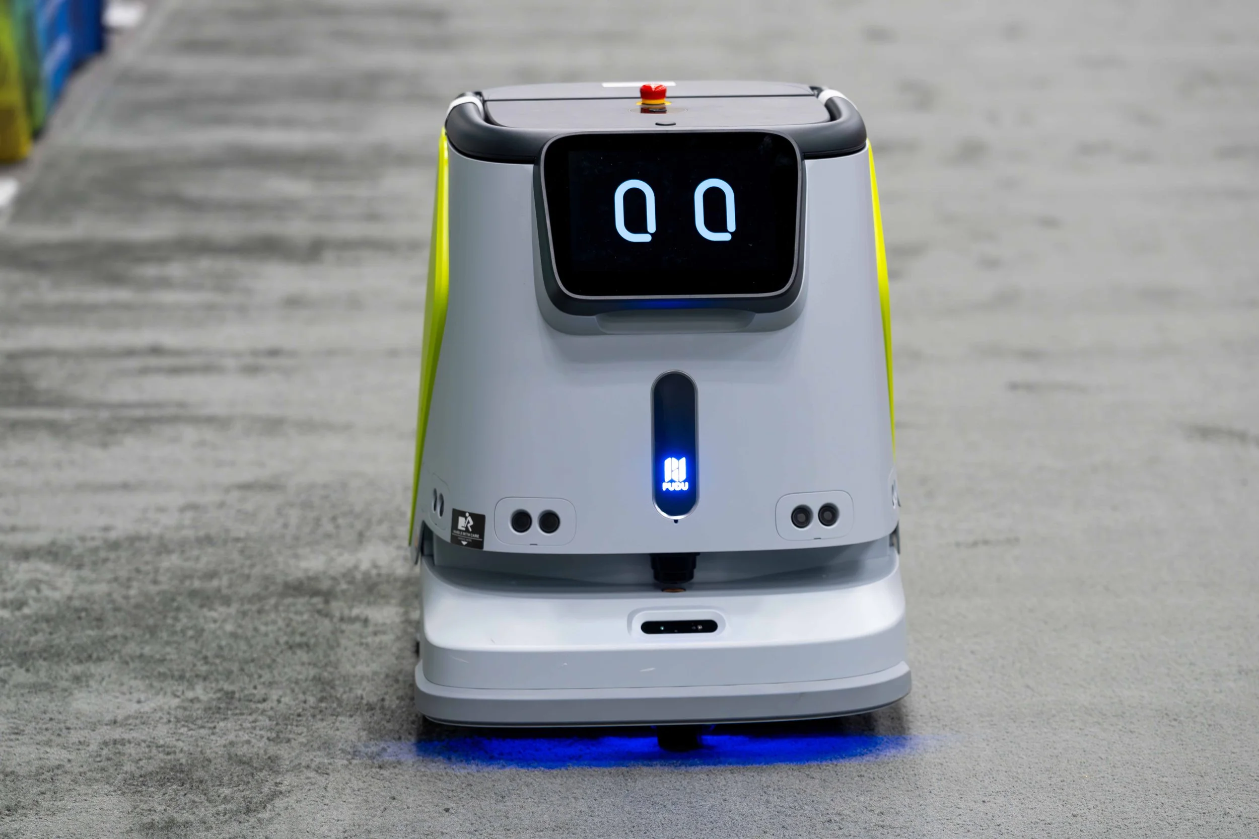 A delivery robot on a concrete surface with digital eyes displayed on its screen, a blue logo on its body, and blue lights shining beneath it.
