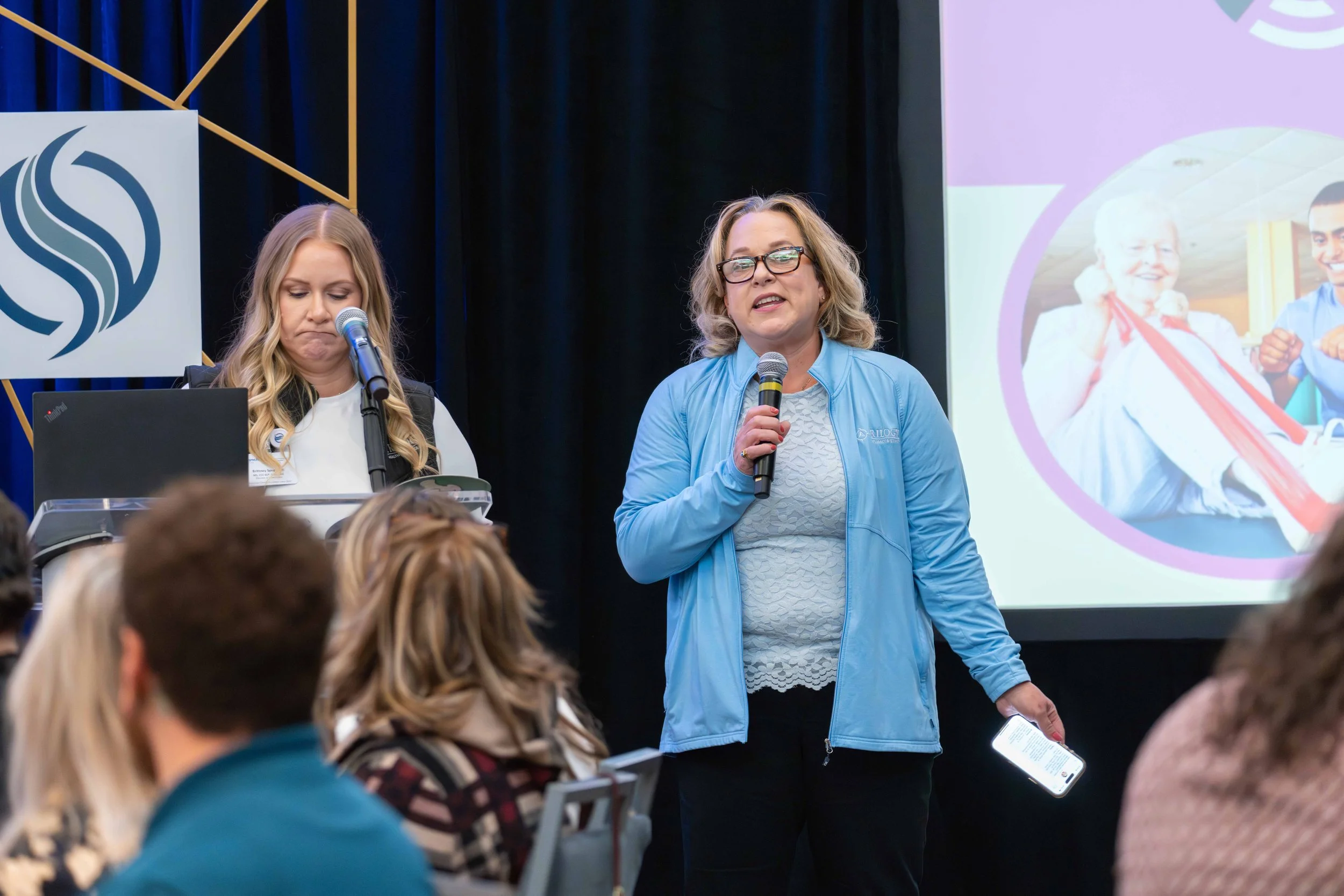 Two women speaking at a conference or seminar, one holding a microphone and the other working at a laptop, with a projection screen and audience in front.