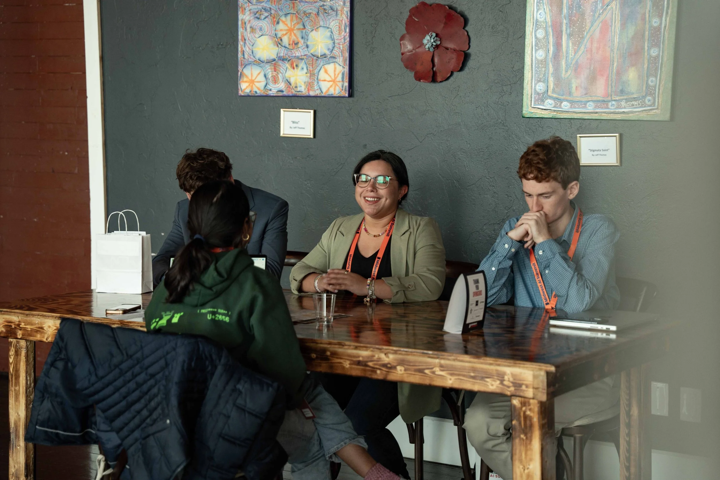 Four people sitting at a wooden table in a casual meeting, with two men and one woman engaging with a woman in a green hoodie. Artwork on the wall behind them.