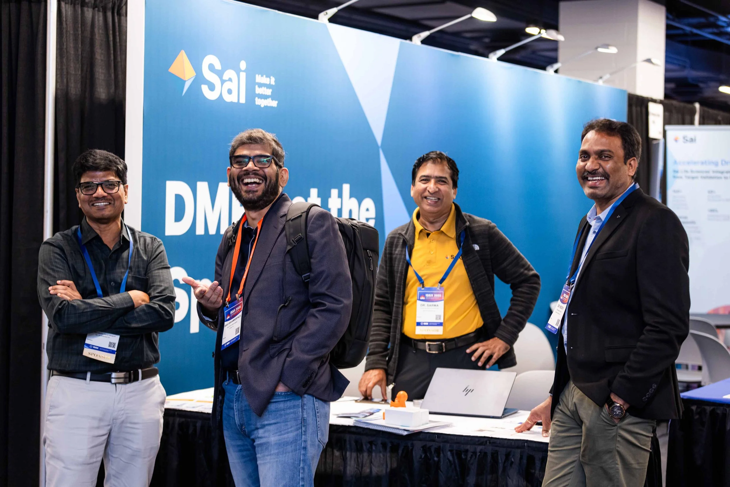 Four men standing together at a conference booth, smiling and posing for the camera. The booth has a blue background with the logos and text related to Sai and DMR.