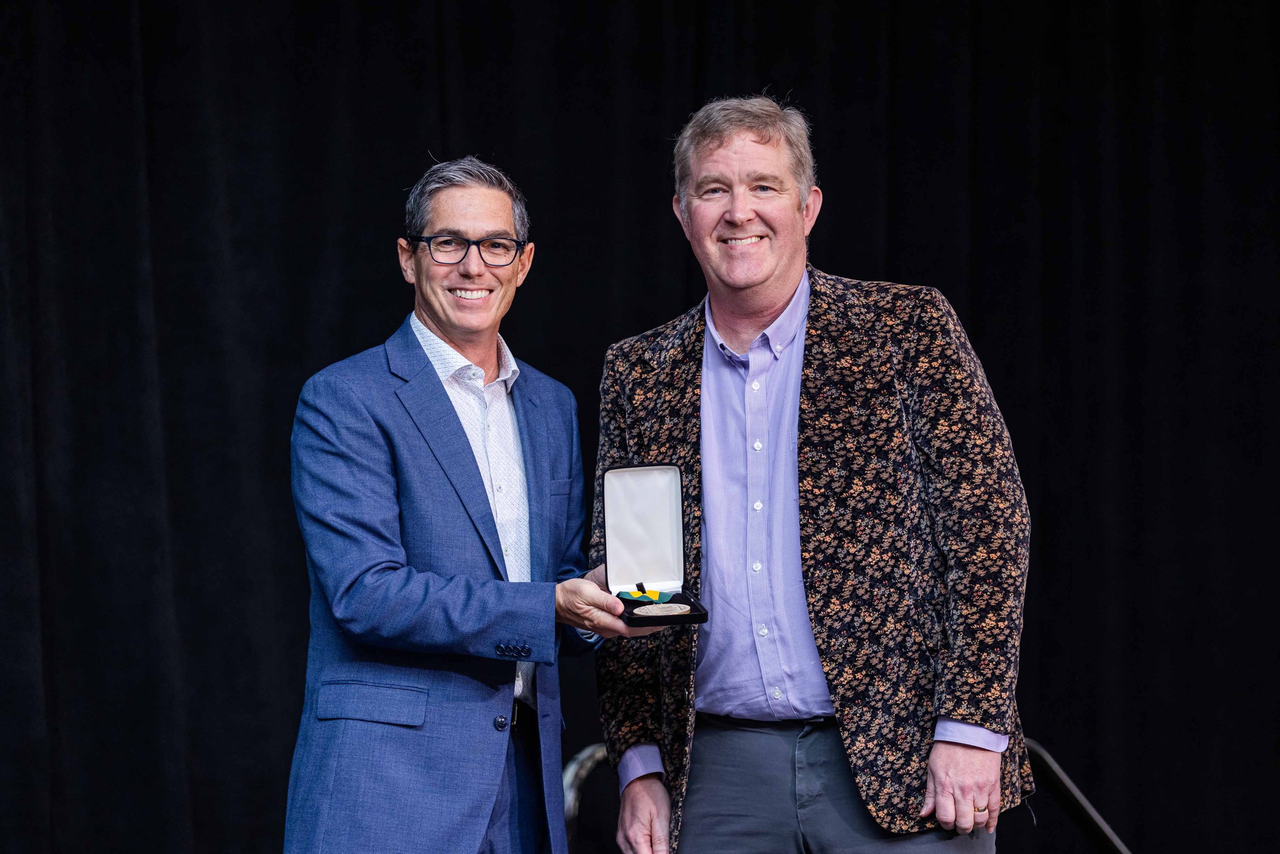 Two men in suits standing on a stage, one is holding a medal in a case, smiling for a photo against a black background.