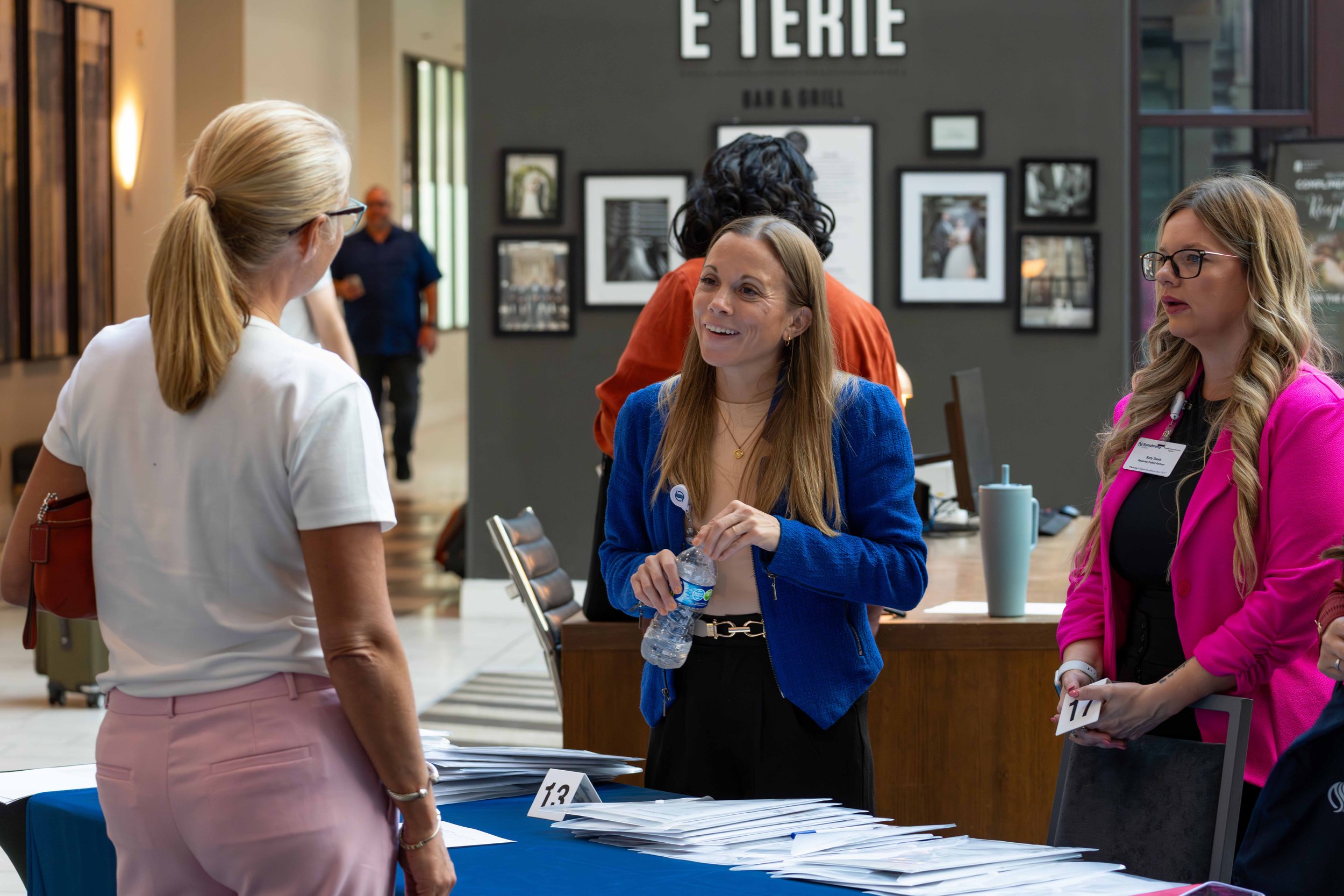 Women are engaged in conversation at a registration or check-in table at an event, with stacks of papers, name tags, and a blue tablecloth.