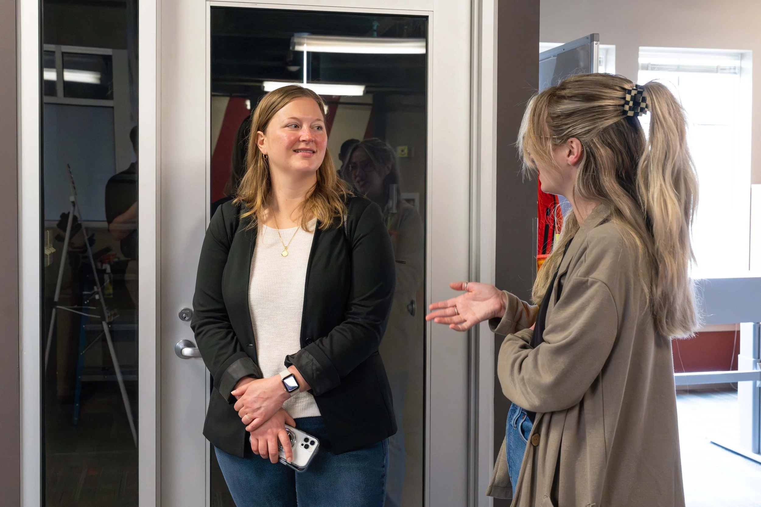 Two women are having a conversation in an indoor setting, standing near a glass door or window. One woman, on the left, is wearing a black blazer over a white top and holding a smartphone. The other woman, on the right, has long blonde hair tied with