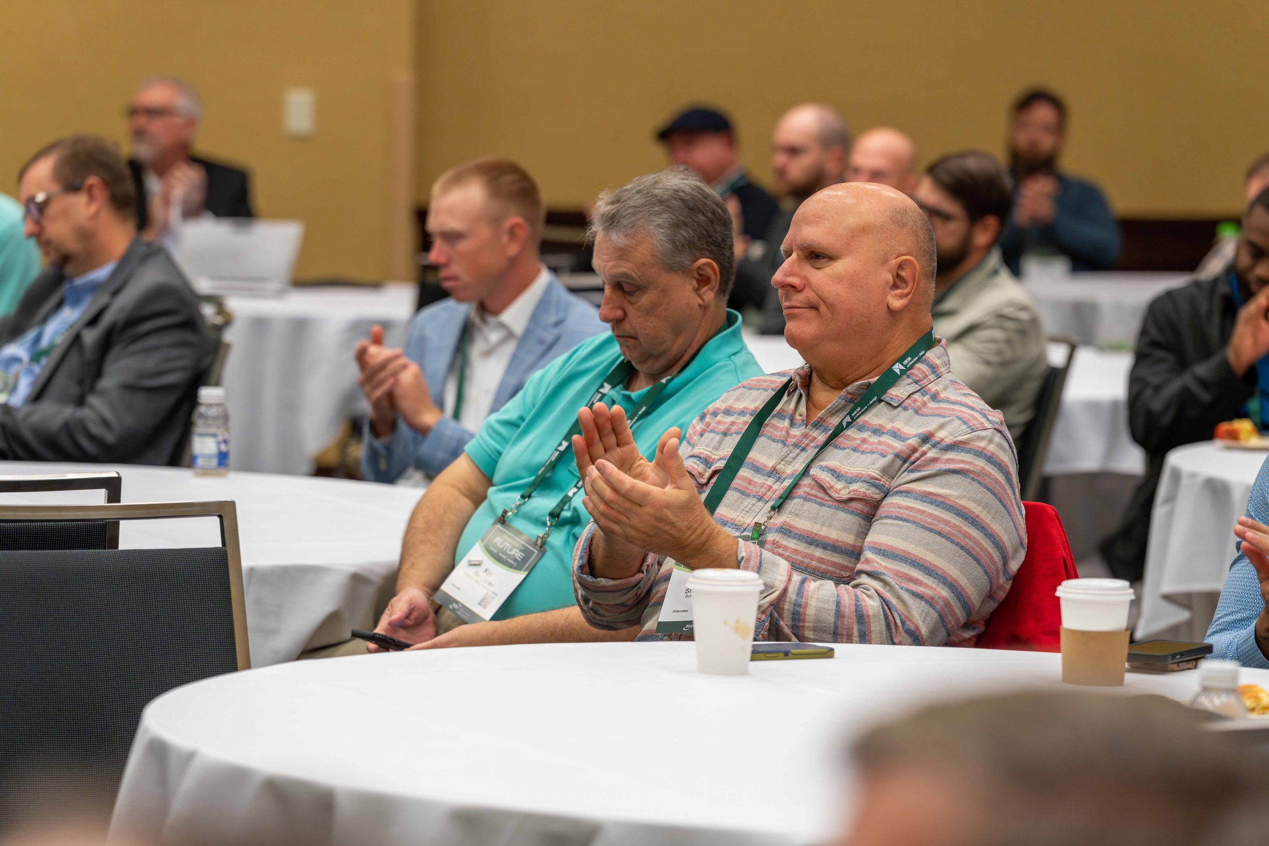 Attendees at a conference sitting at round tables, some clapping and listening attentively, with conference badges around their necks.