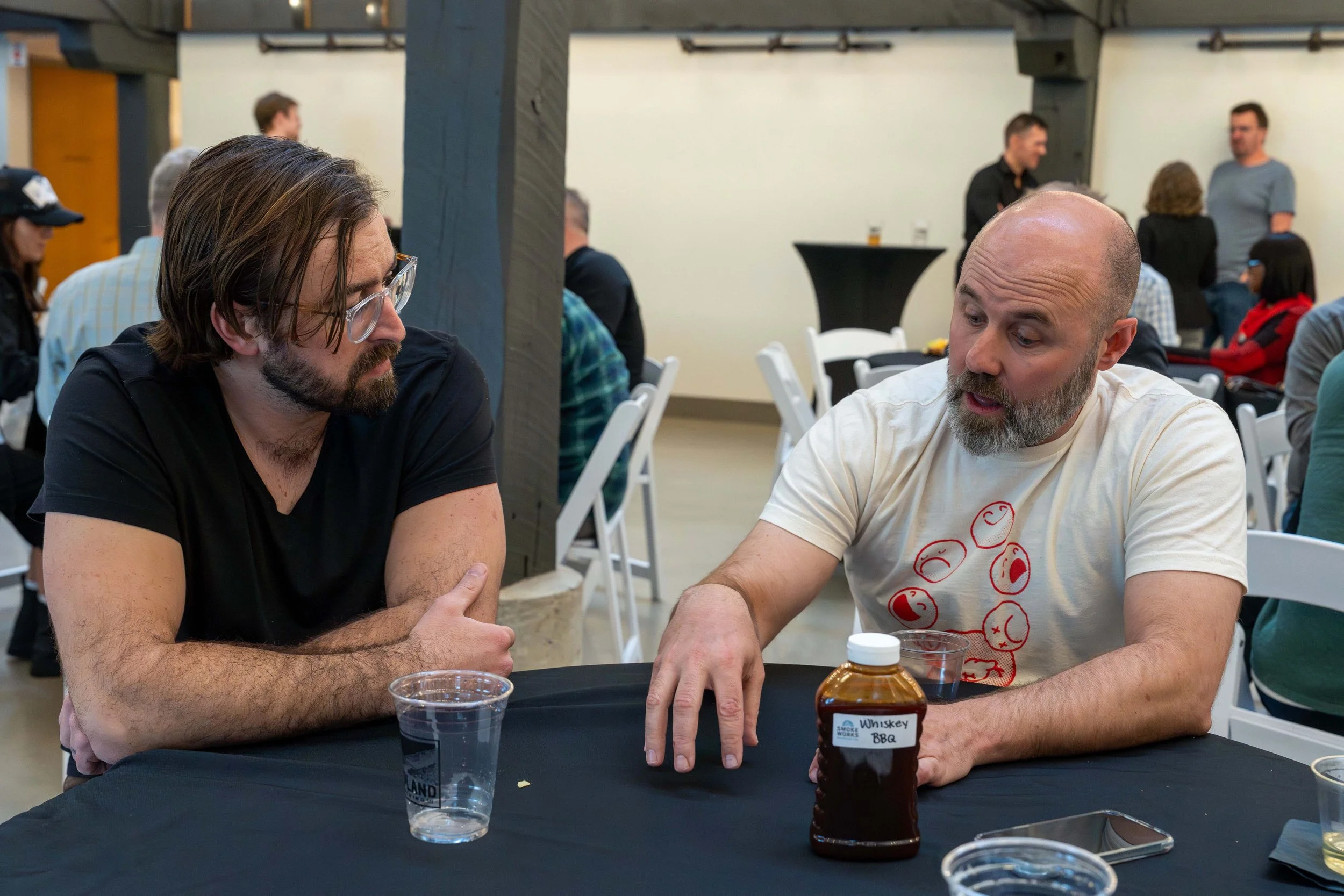 Two men sitting at a black-covered table, engaged in conversation. The man on the right, with a shaved head and gray beard, is gesturing toward a bottle labeled 'Whiskey BBQ'. The man on the left, with long brown hair, glasses, and a beard, is listen