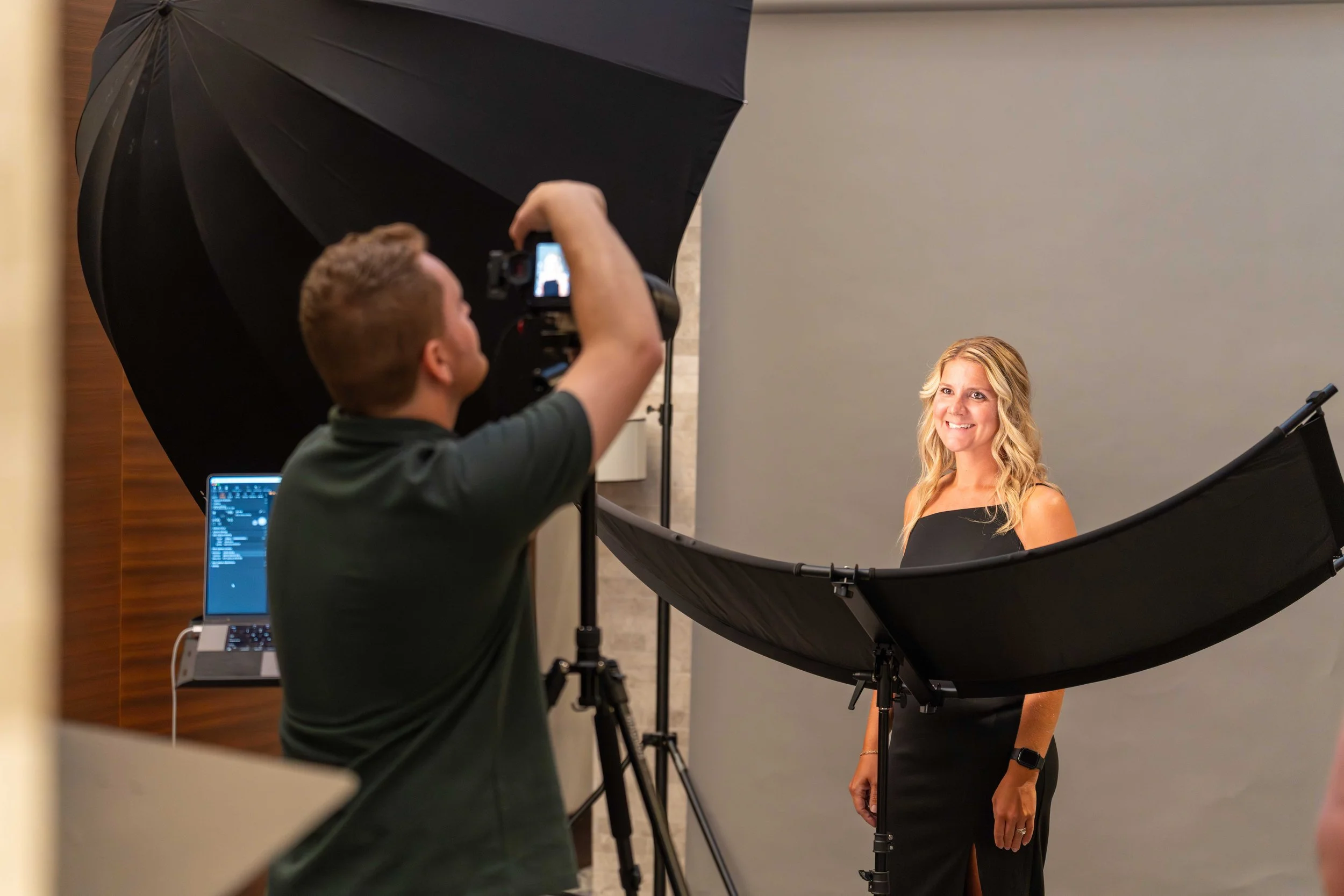 A woman in a black dress poses for a photoshoot with a gray backdrop, while a photographer takes her picture using professional lighting equipment and a camera on a tripod.