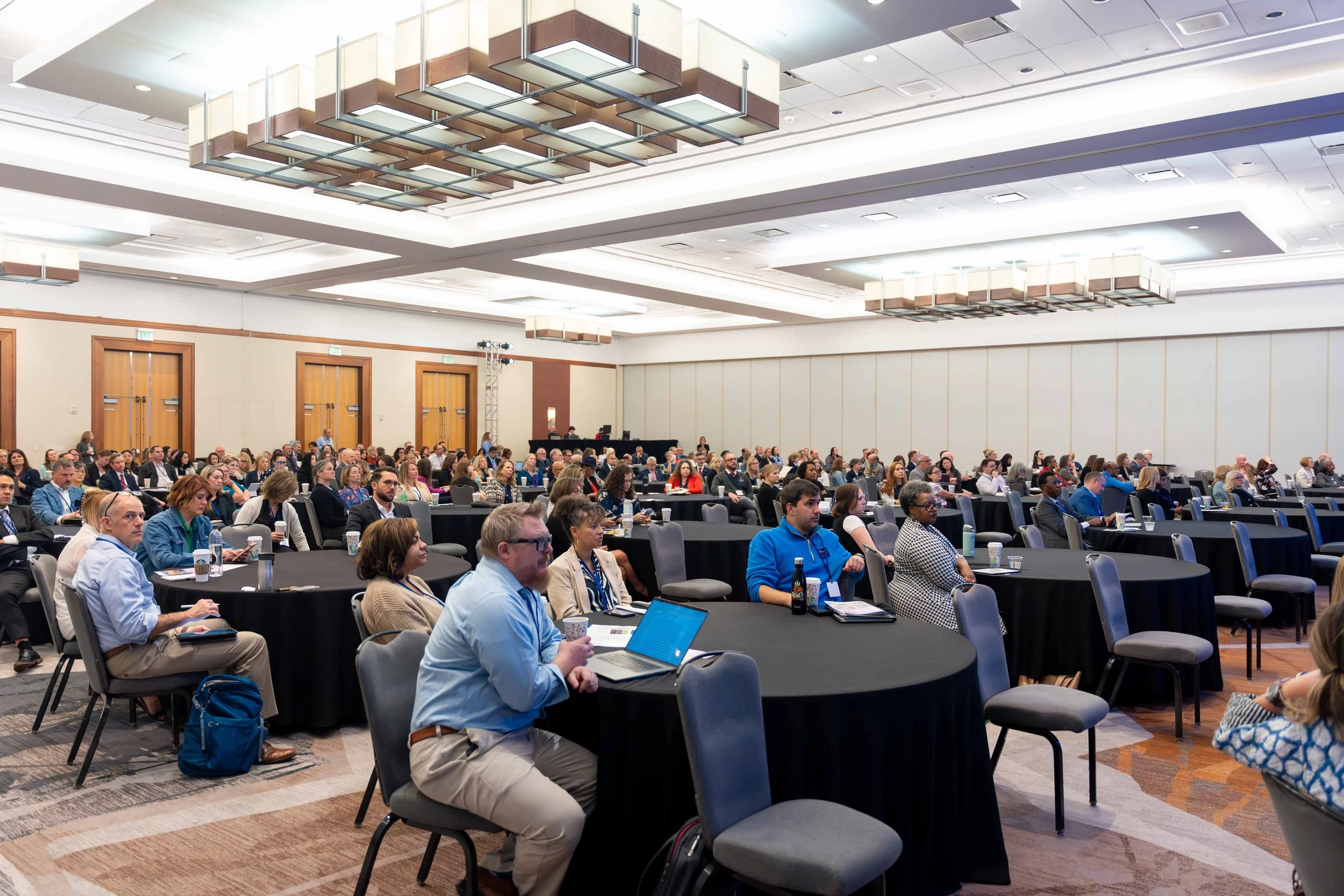 A large conference room filled with audience members sitting at round tables, listening to a presentation. The room has modern lighting and wood-paneled walls.