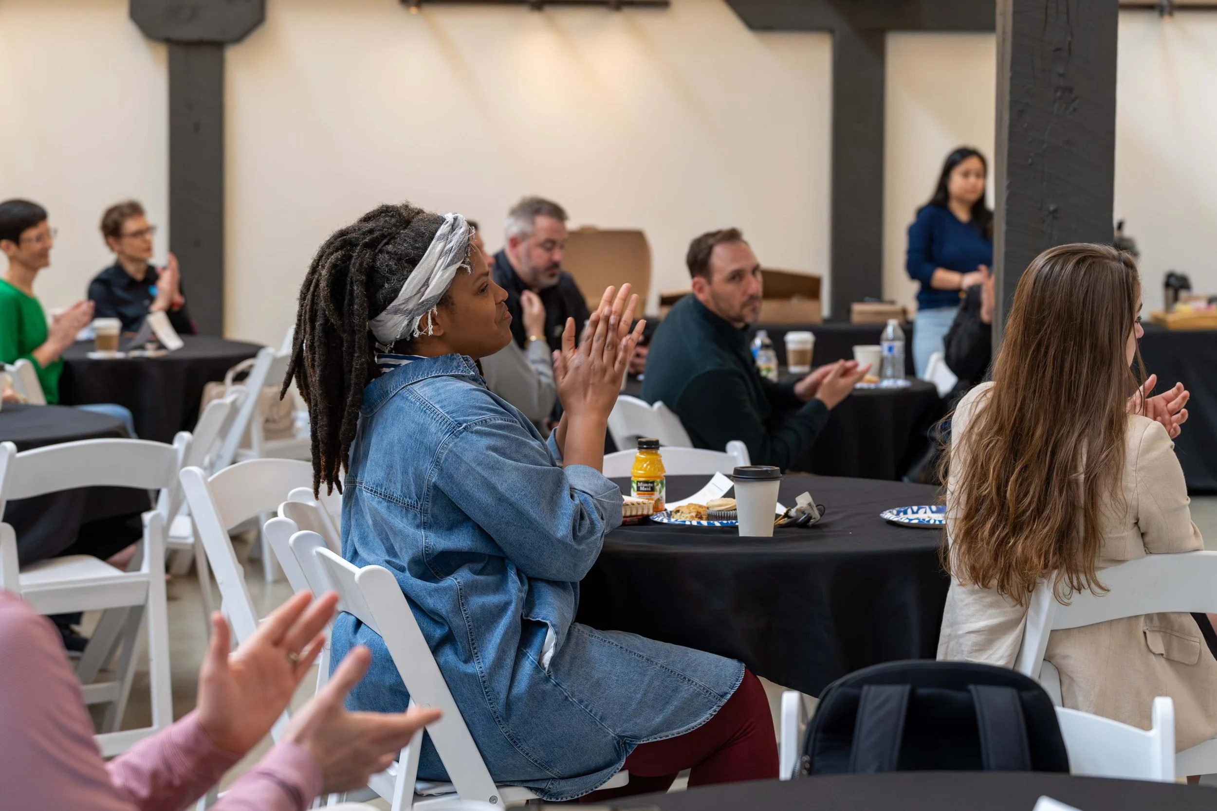 A group of people seated at tables during a meeting or conference, some clapping and others listening attentively.