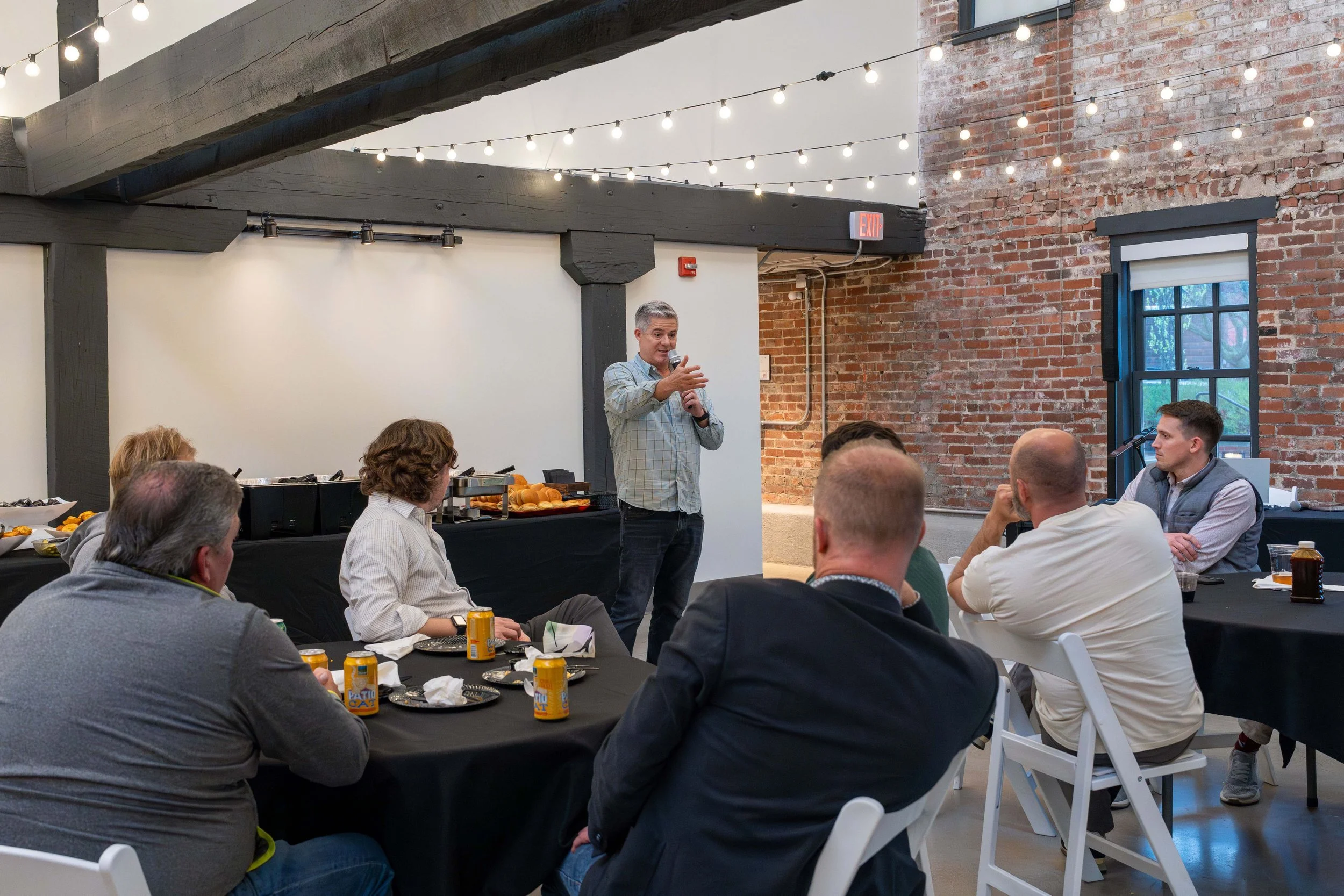 A man is giving a speech to a seated audience in a rustic-style event space with brick walls and string lights, while holding a microphone and gesturing with his hand.