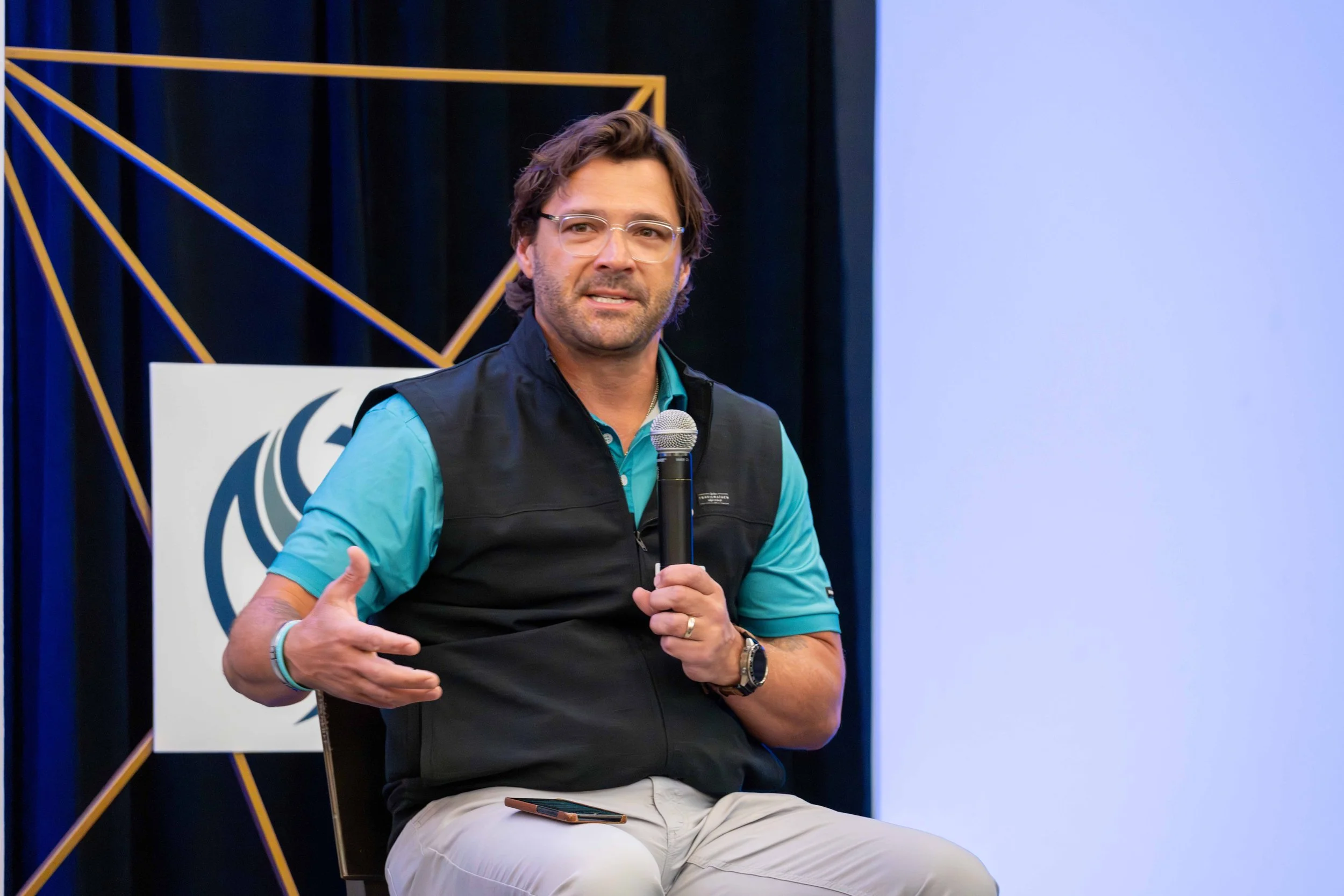 A man with glasses and a beard holding a microphone, sitting on a chair during a presentation or discussion.