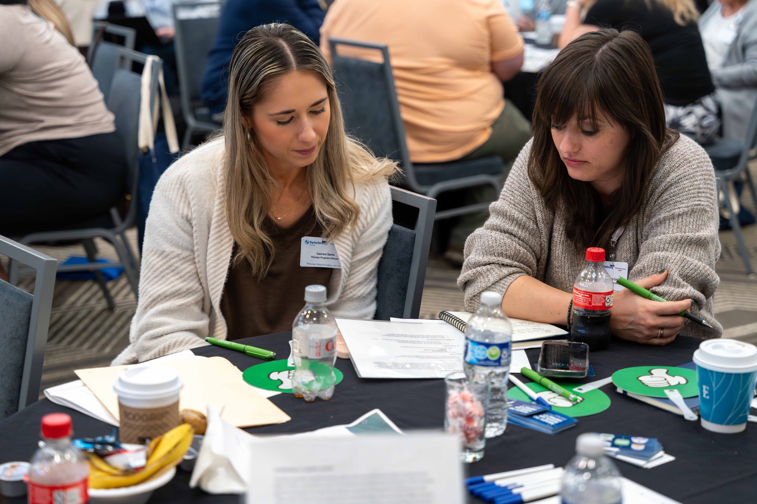 Two women sitting at a table during a conference or workshop, with papers, pens, water bottles, coffee cups, and snacks on the table.