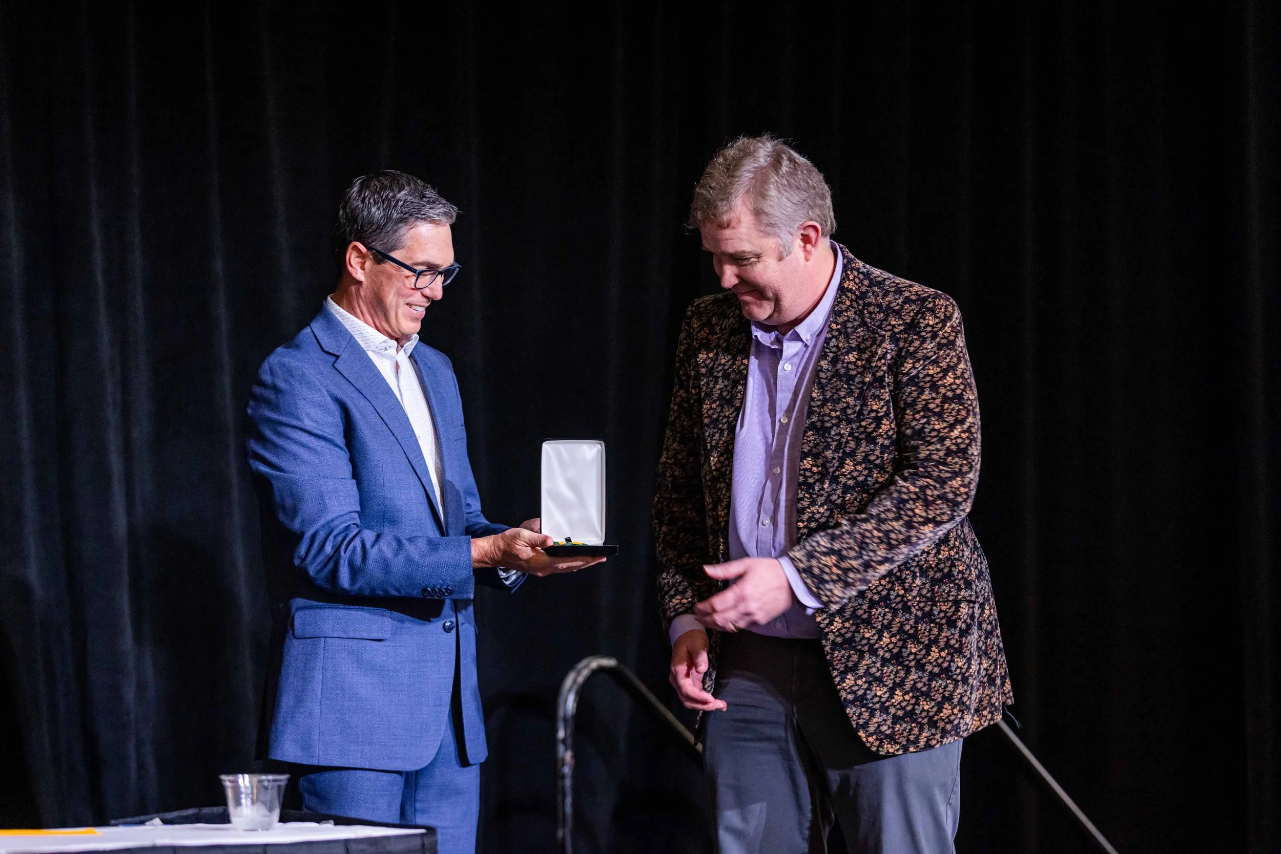 Two men in suits standing on stage, one handing a medal or award to the other, during an award ceremony or presentation.