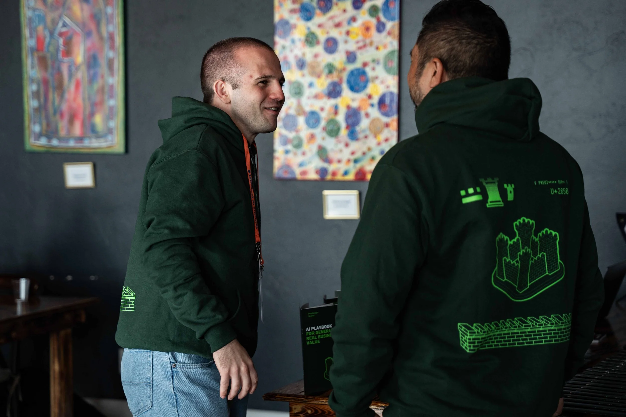 Two men wearing green hoodies and jeans engaging in conversation at an art gallery.
